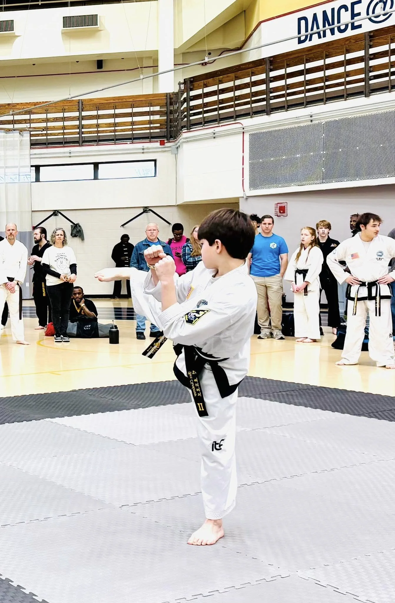 A young martial artist practicing a kick in a gym during a martial arts event, with spectators and fellow practitioners watching.