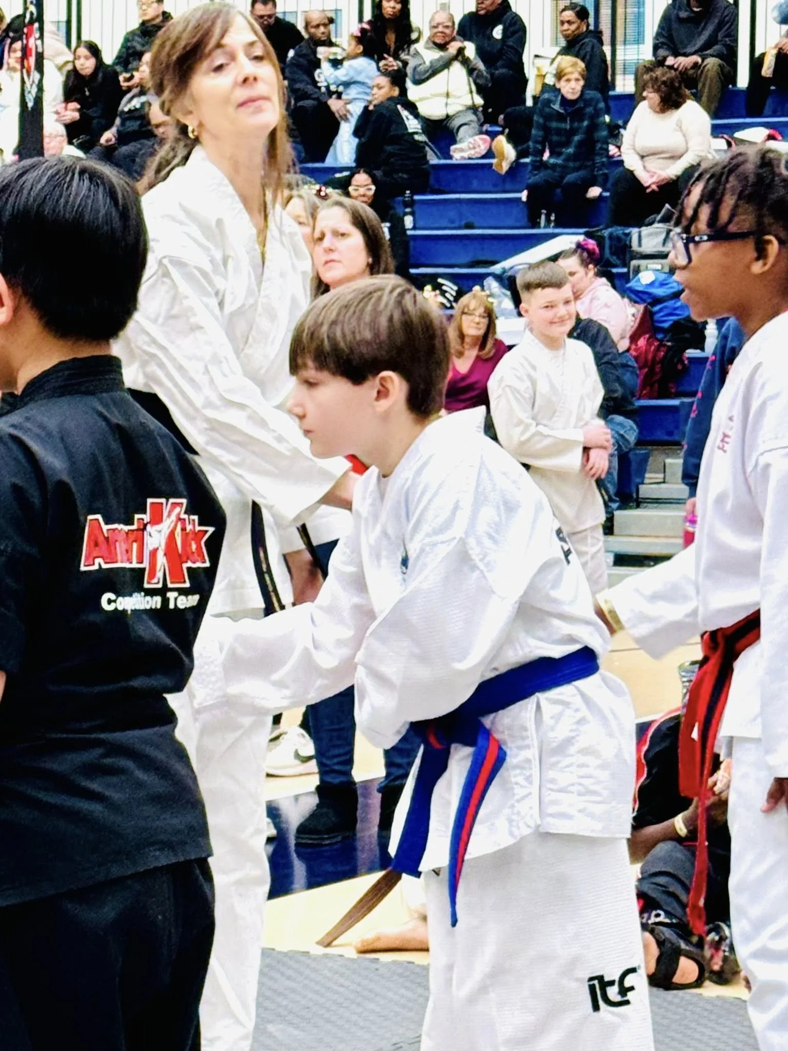 Martial arts competition with children in uniforms, some shaking hands, in an indoor arena with spectators watching.