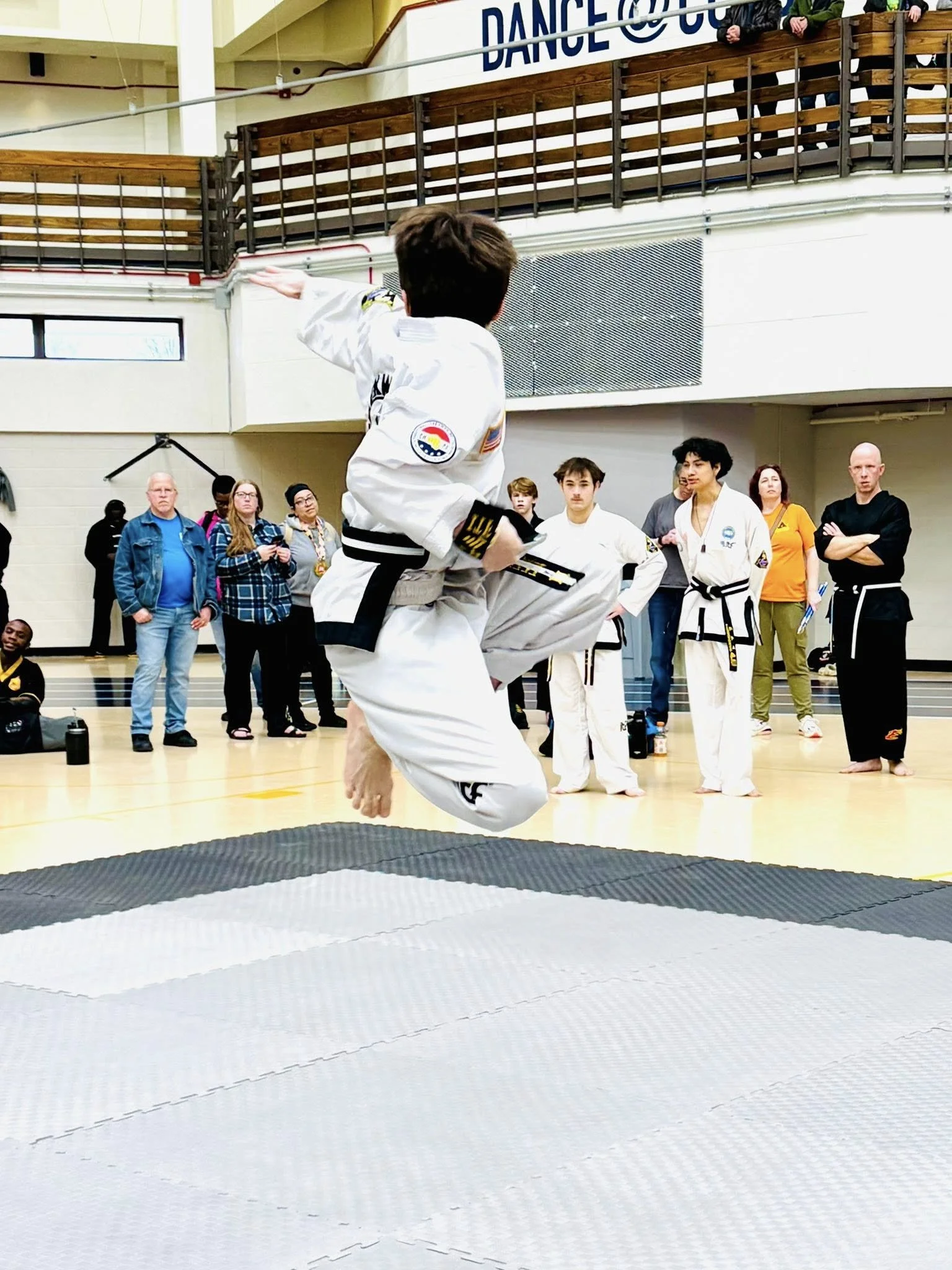 A person in a martial arts uniform performs a jump kick during a martial arts demonstration in a gymnasium, with spectators and fellow martial artists watching.