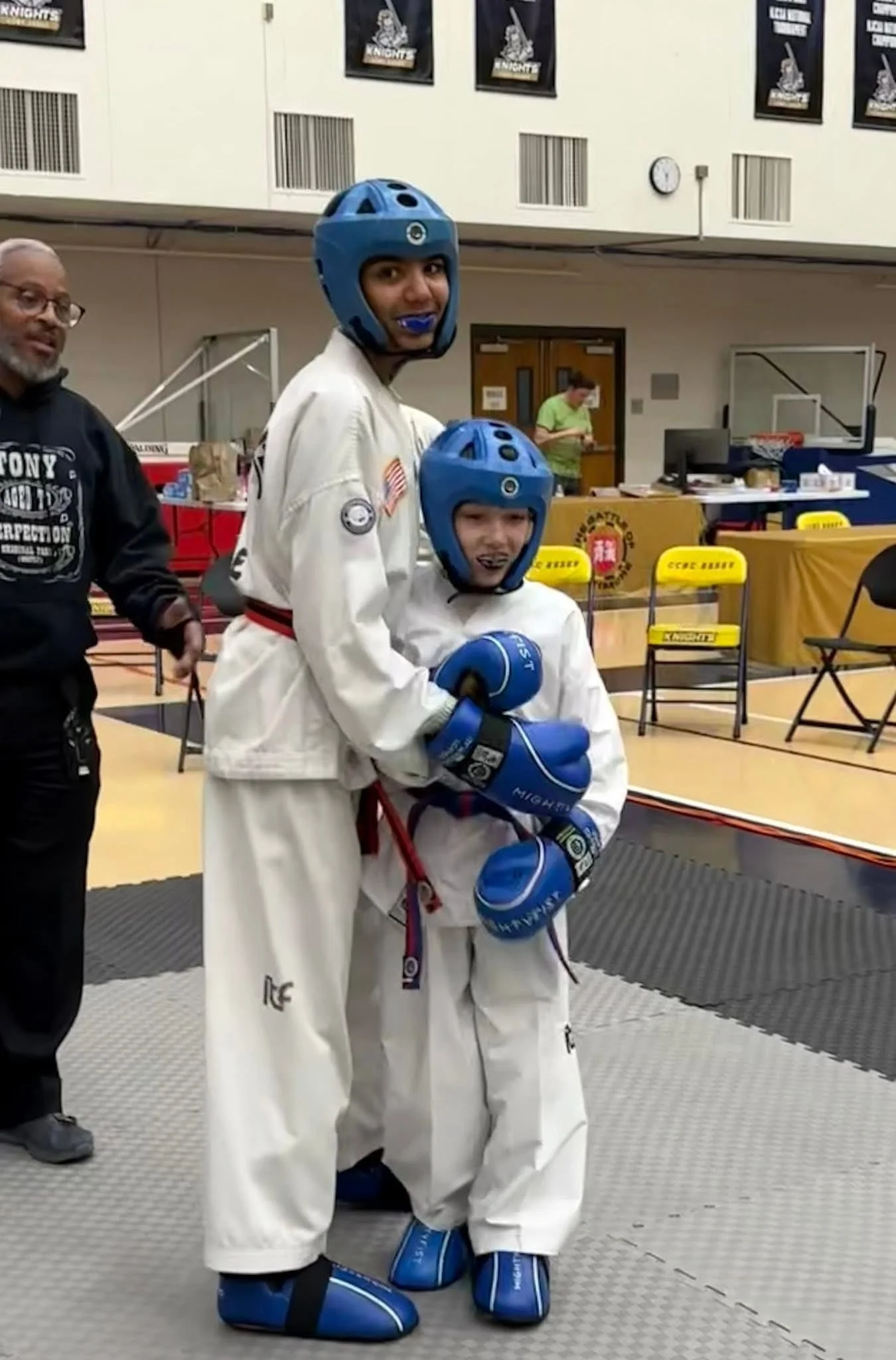 Two children in martial arts uniforms with blue protective gear, posing in a gymnasium, with a man and a woman in the background.
