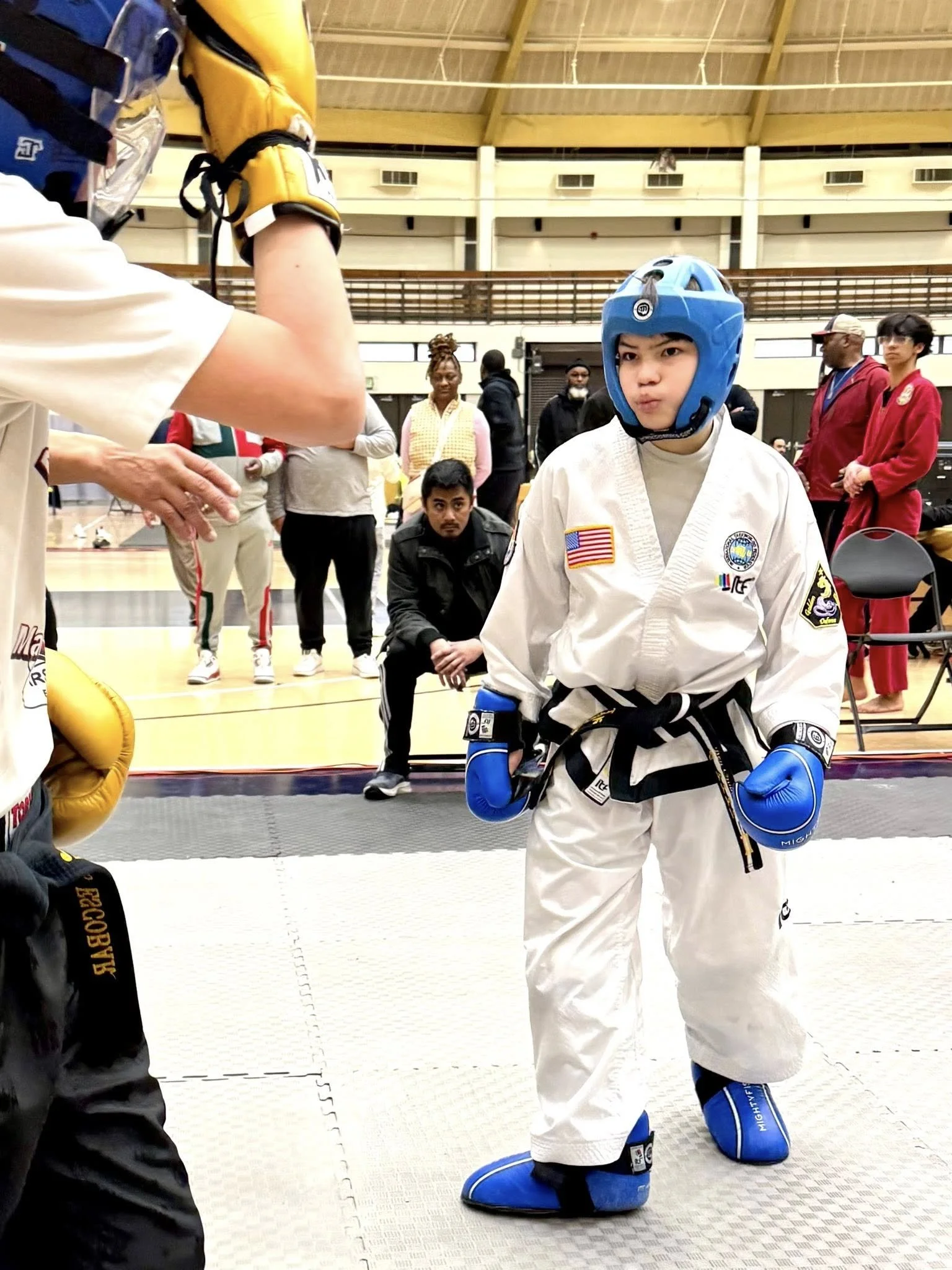 Young martial artist in a white karate gi with patches, wearing blue protective gear including a helmet, gloves, and foot guards, standing on a training mat in a gymnasium, preparing for a match.
