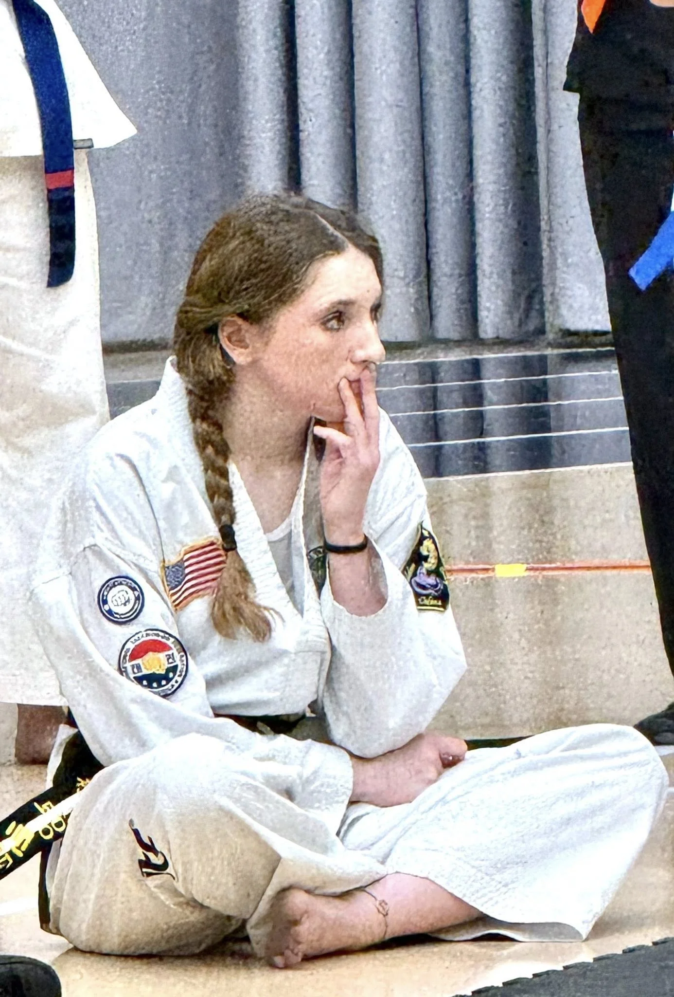 A woman sitting on the floor in a martial arts uniform, with patches on her sleeve, her hair in a braid, in a contemplative pose with her fingers near her mouth, in front of a curtain.