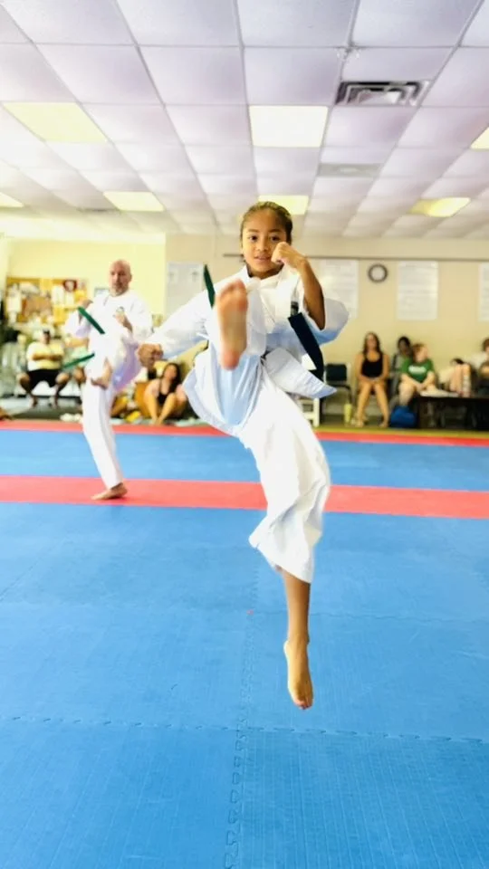 Young girl performing a high kick during a martial arts demonstration in a dojo, with spectators seated in the background.
