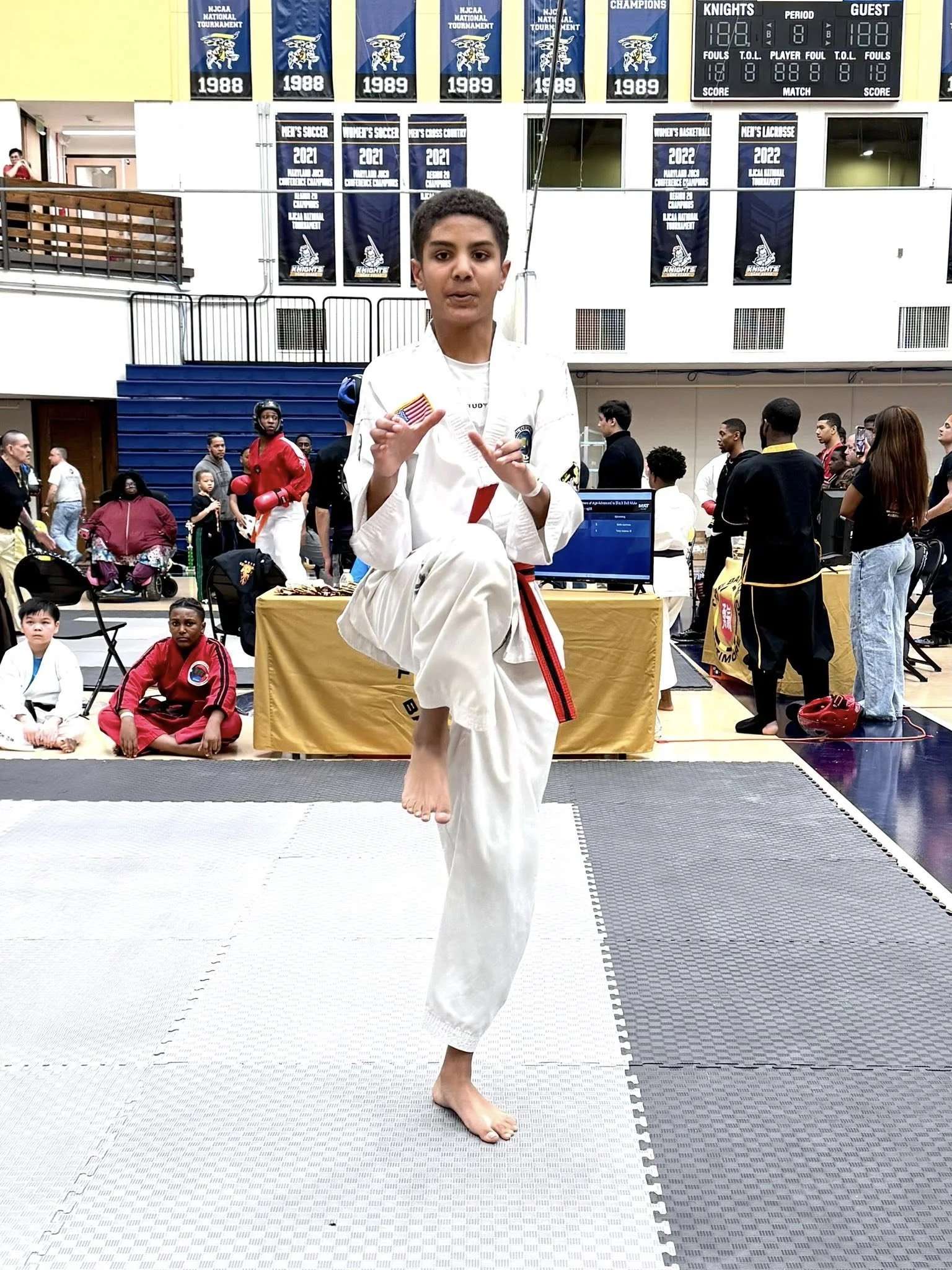 Young martial artist in a white gi demonstrates a kick at a martial arts event in a gymnasium.
