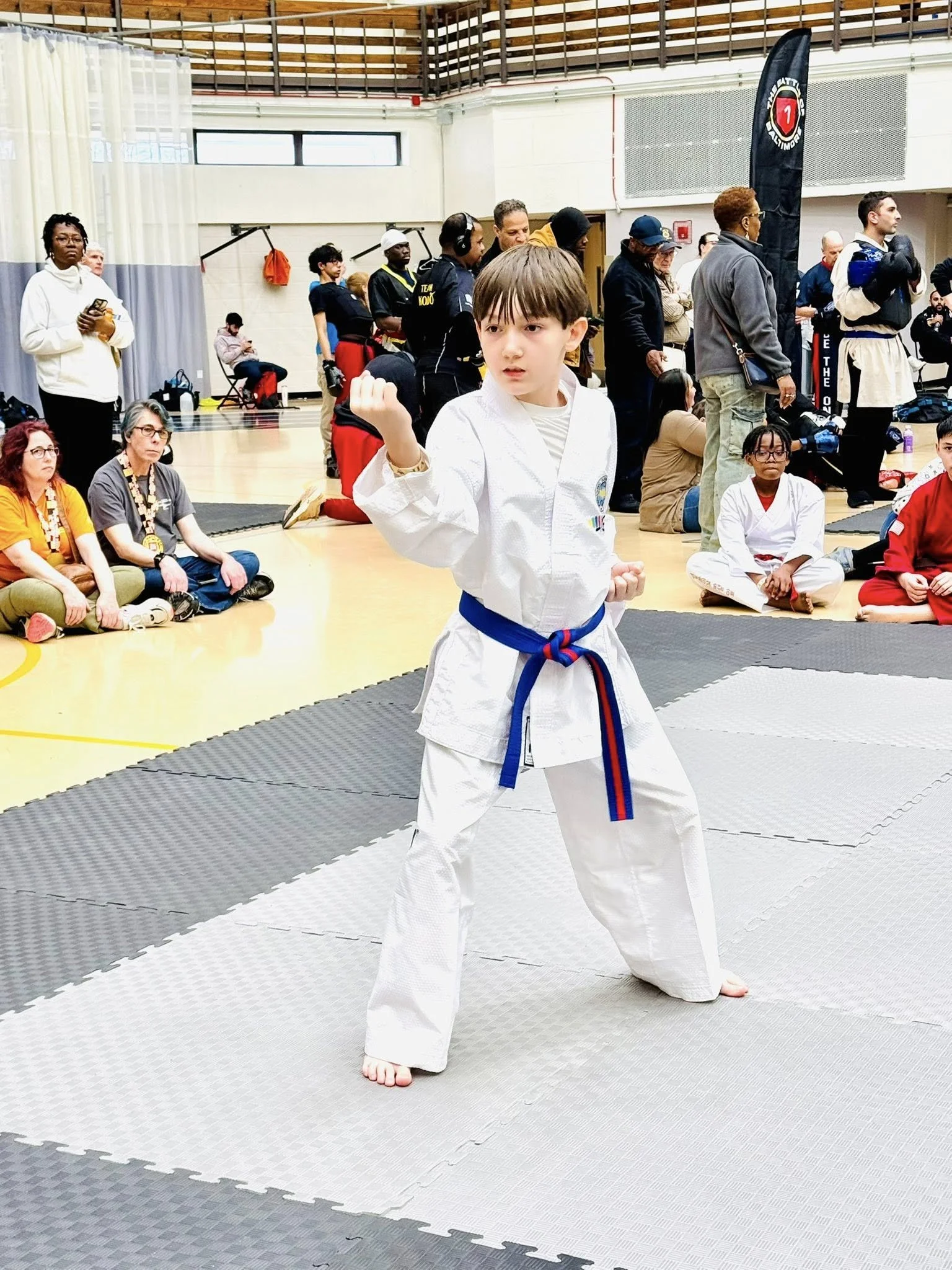 Young boy in a white martial arts uniform with a blue belt performing a move on a matted competition area at a martial arts tournament, with spectators and other competitors in the background.