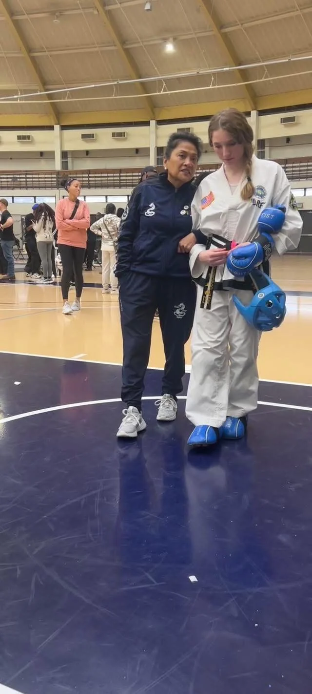 A young female martial artist in a white gi and blue protective gear being assisted by an older woman in a navy tracksuit inside a gym with a high ceiling. Several people are in the background.
