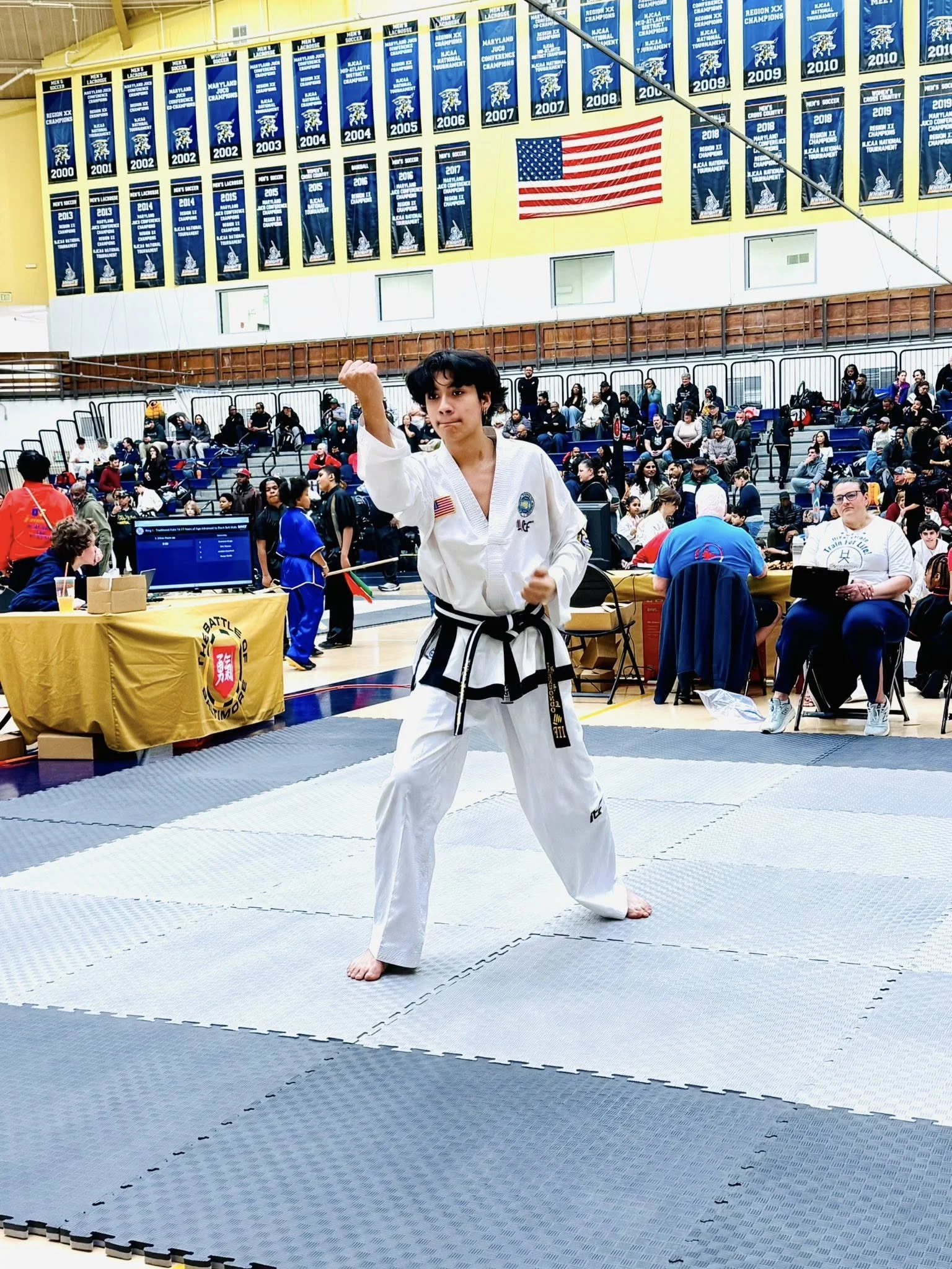 A young martial artist in a white uniform with a black belt, performing a martial arts stance at a competition in a gymnasium filled with spectators, banners, and flags on the wall.