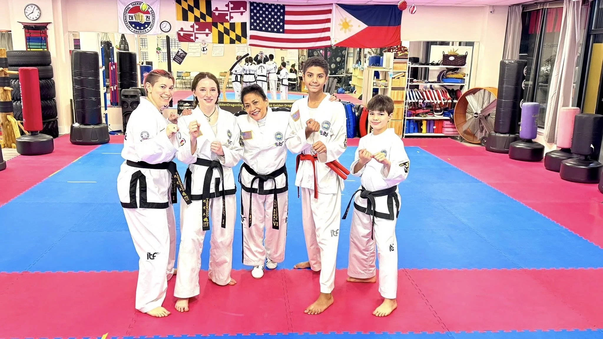 Group of five people in martial arts uniforms with black belts, standing on a martial arts training mat inside a dojo, smiling at the camera. Flags and martial arts equipment are visible in the background.