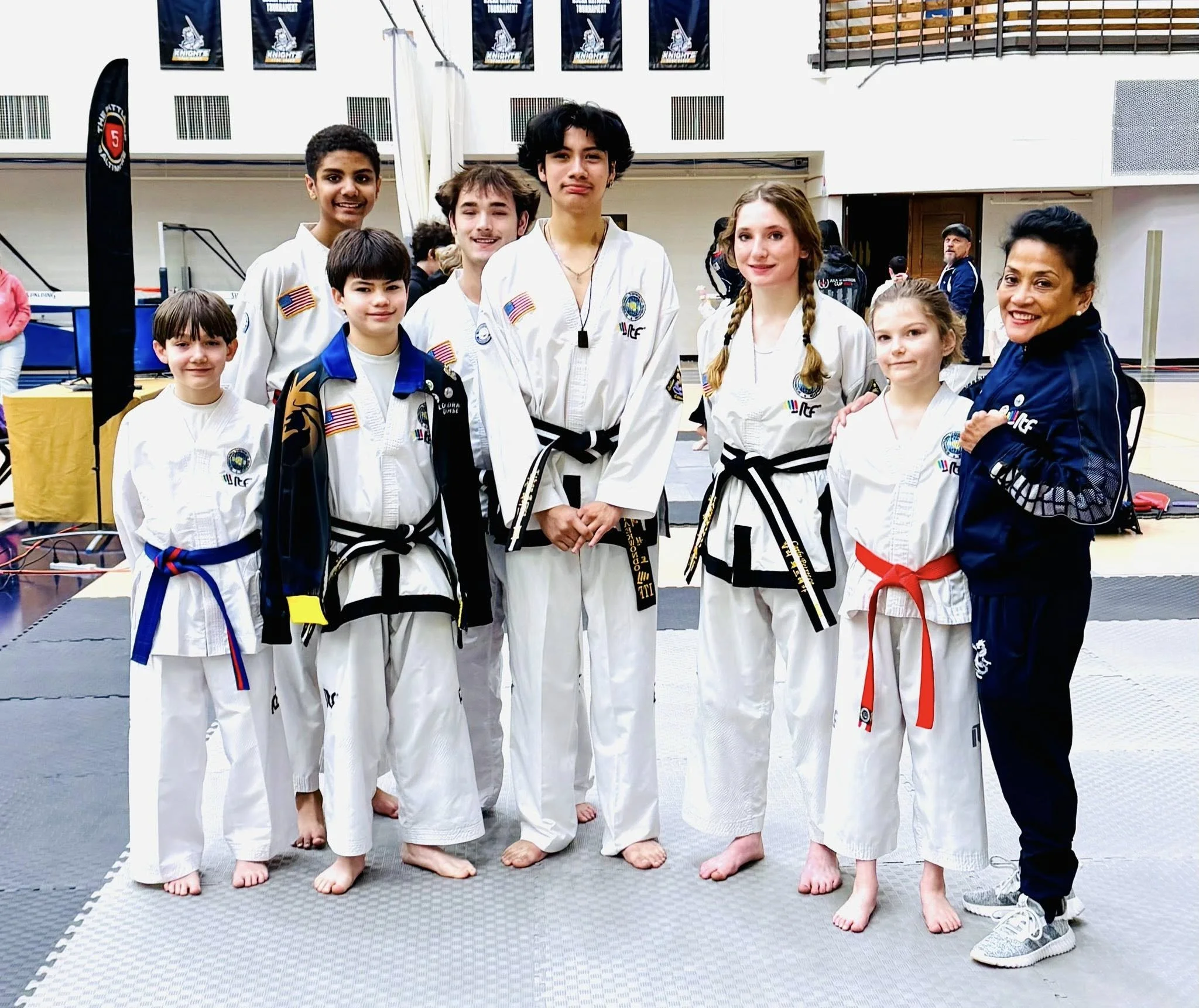 Group of nine young martial artists and a female coach posing in a gym, wearing doboks with various colored belts, after a tournament.