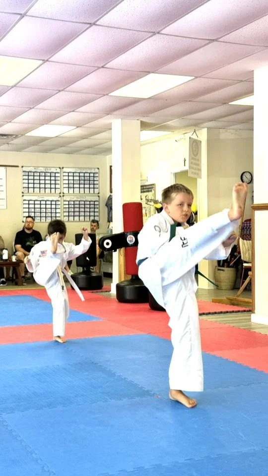Two children practicing martial arts in a dojo, performing high kicks, with spectators seated in the background.