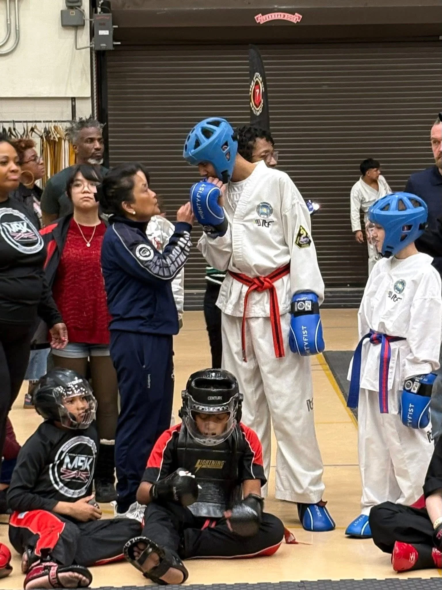 A group of martial artists, some wearing white uniforms with colored belts and helmets, standing and sitting in a gym, with some people watching in the background.