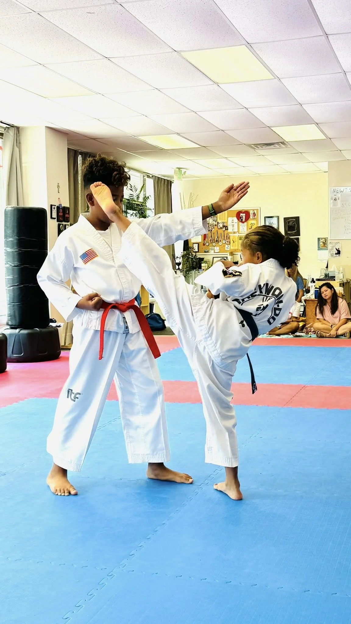 Two children practicing martial arts in a dojo, wearing white gis and black belts, with one performing a kick while the other blocks.