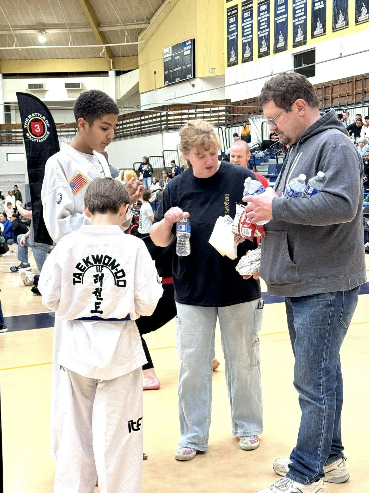 Two children in taekwondo uniforms talking with two adults in a gymnasium, with spectators in the background and banners hanging from the ceiling.