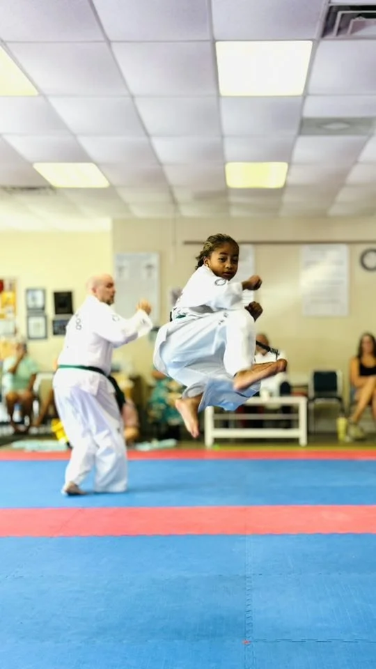 Young girl performing a high jump during a martial arts class in a gym, with a man in a martial arts uniform in the background.