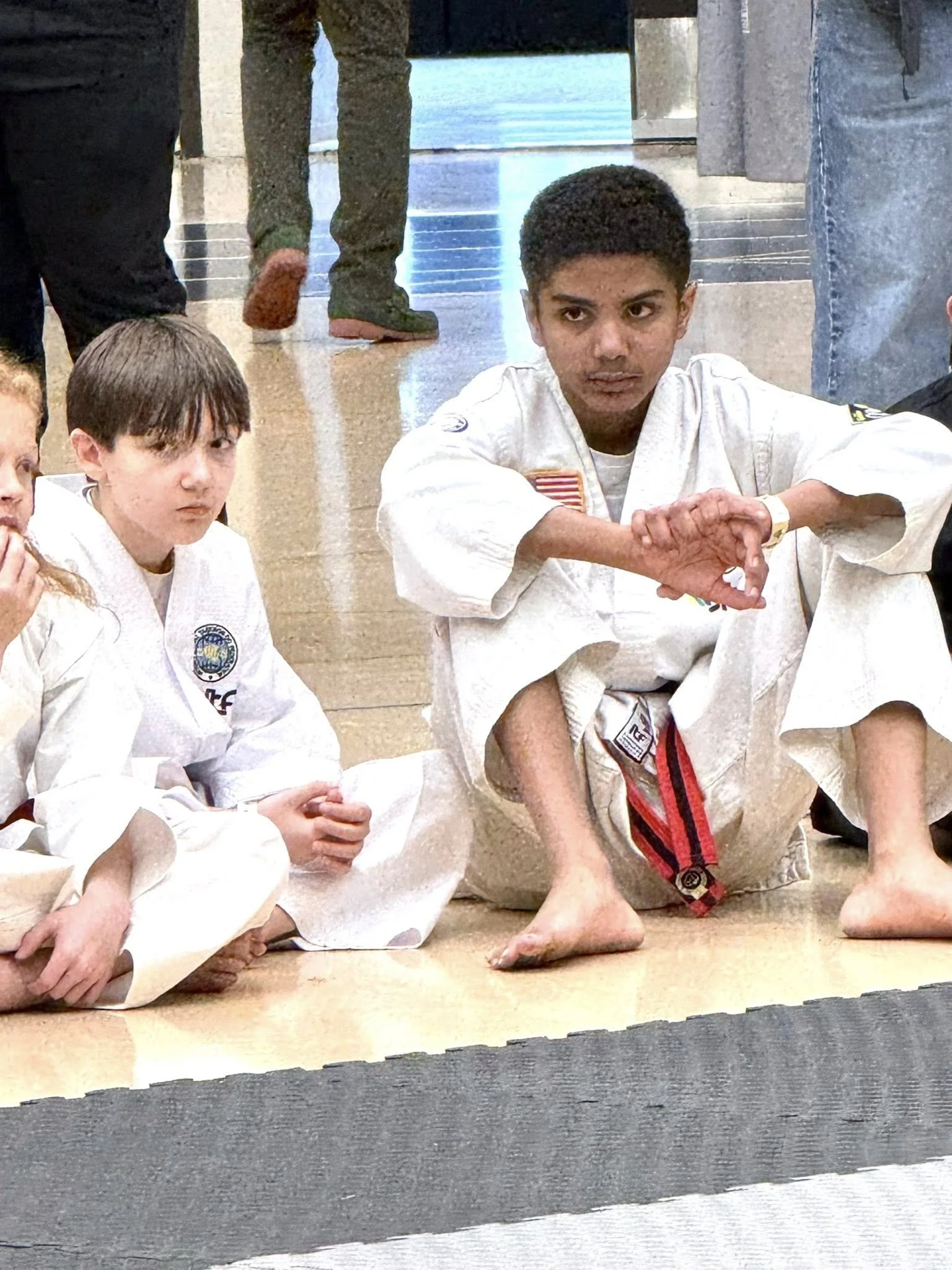 Group of children in martial arts uniforms sitting on the floor during a class or demonstration, with a boy in the center making a hand gesture.