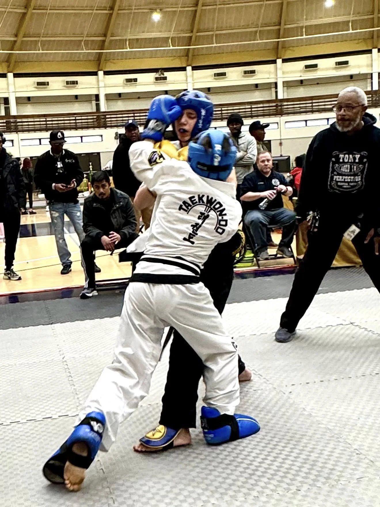 Two martial artists competing in a taekwondo match, both wearing protective gear including helmets, gloves, and foot guards. The competitor in white has their back to the camera, while the other is facing forward. A referee stands nearby, and spectat