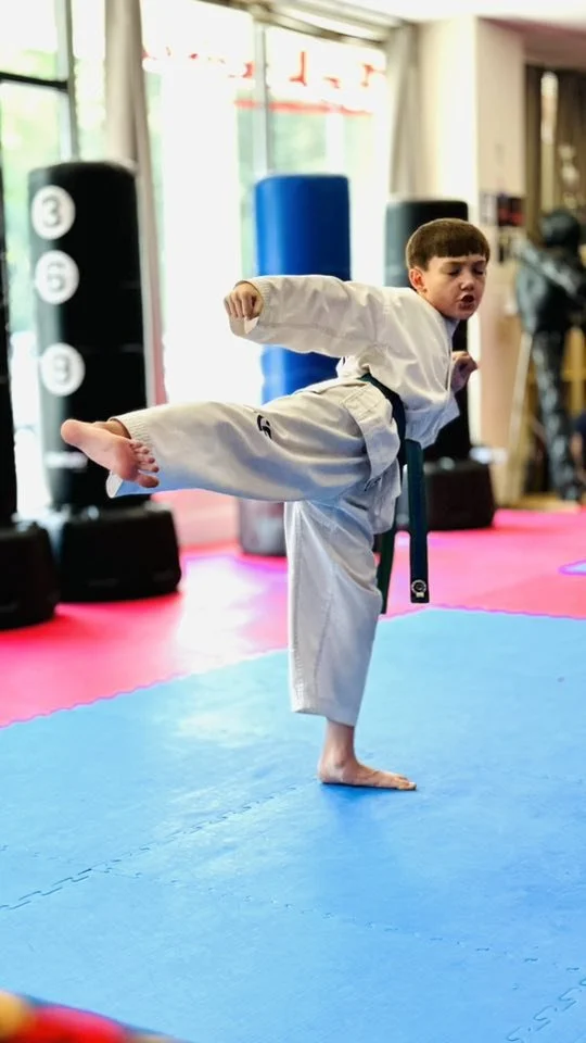 Young boy practicing karate in a martial arts gym, executing a high kick while wearing a karate gi on a blue mat.