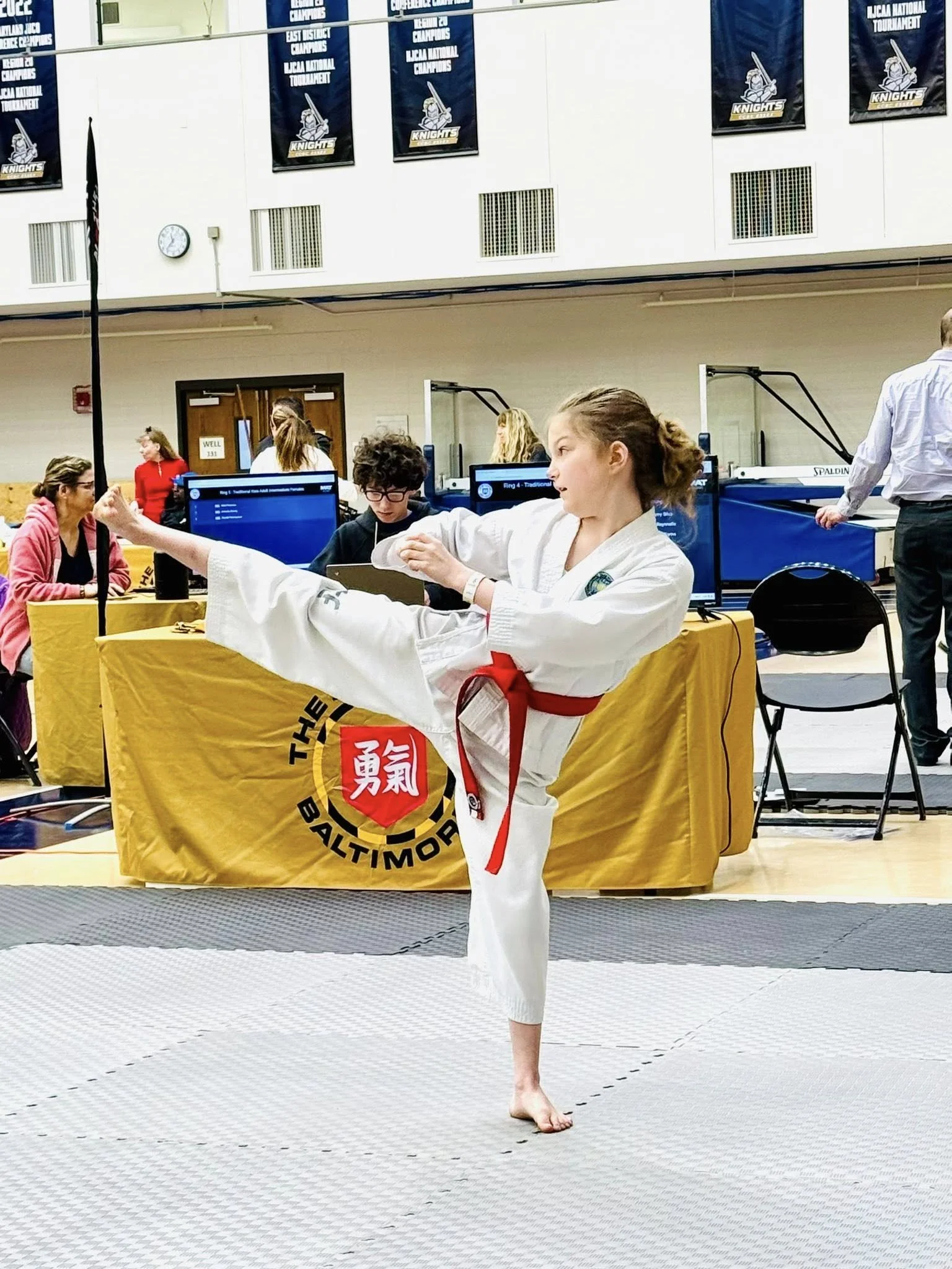A young female martial artist in a white karate gi with a red belt is performing a high kick during a martial arts competition, with spectators and officials in the background at an indoor sports facility.