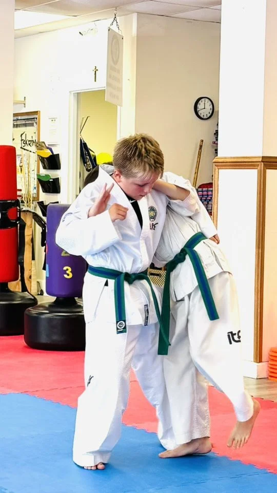 Two young children practicing martial arts wearing white gi uniforms with green belts, one child demonstrating a technique while the other child is being helped or guided in a dojo with martial arts equipment in the background.