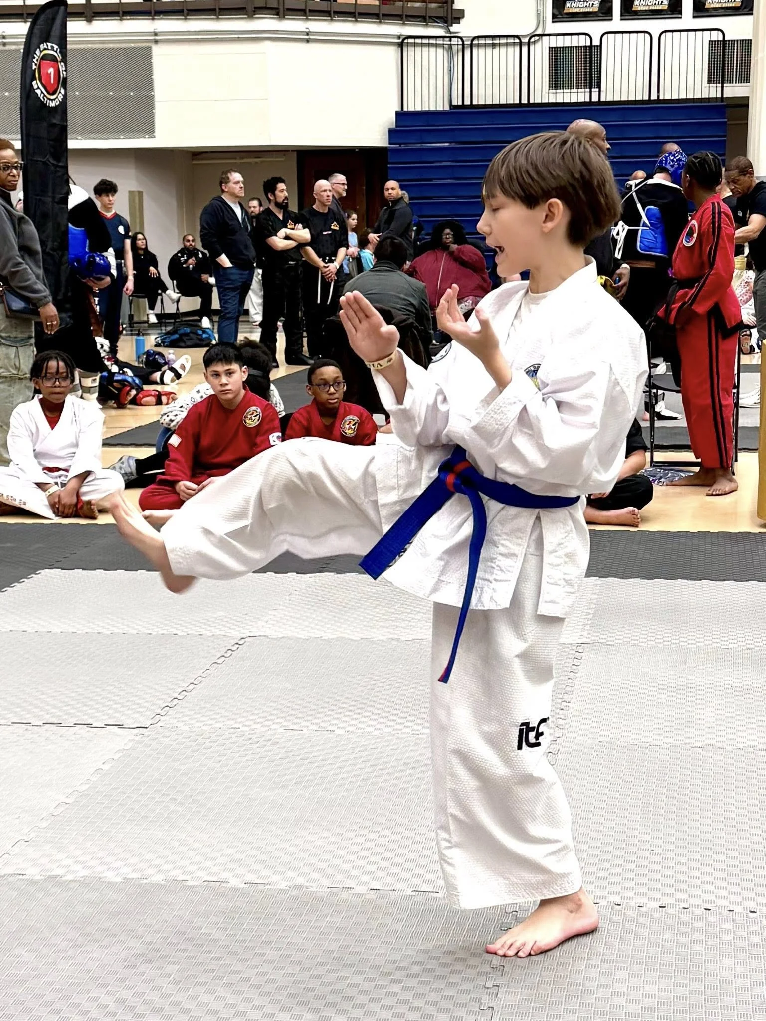 A young girl in a white karate gi and blue belt performing a kick during a martial arts competition on a mat, with spectators and competitors watching in the background.