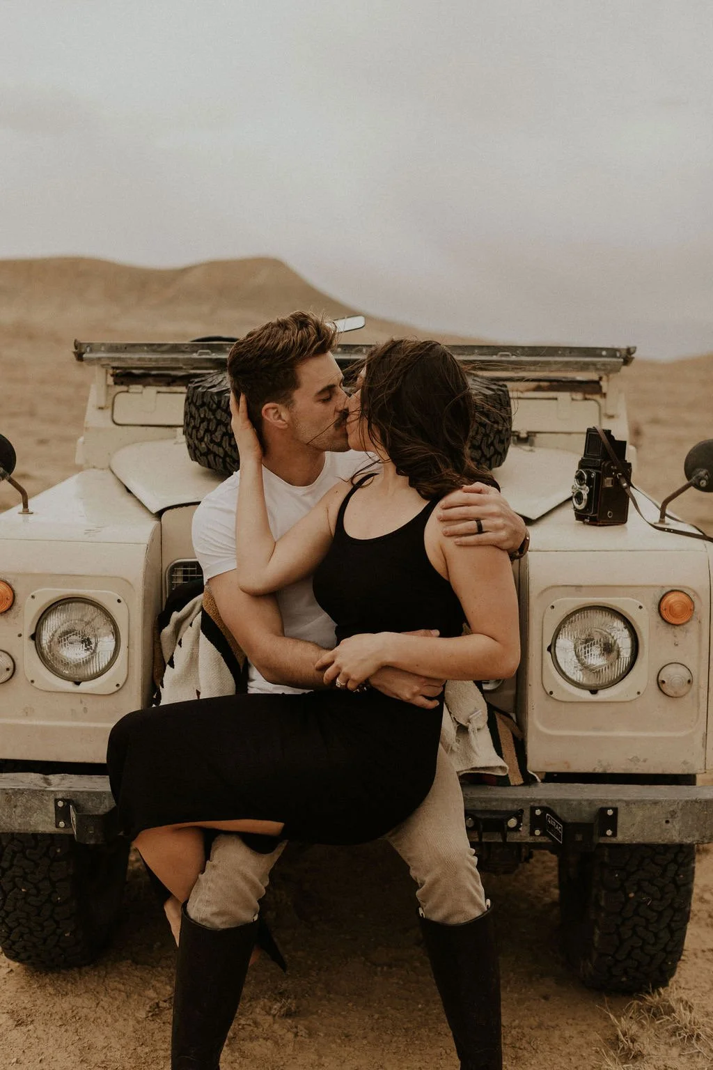 A couple kissing in front of a vintage off-road vehicle in a desert landscape.