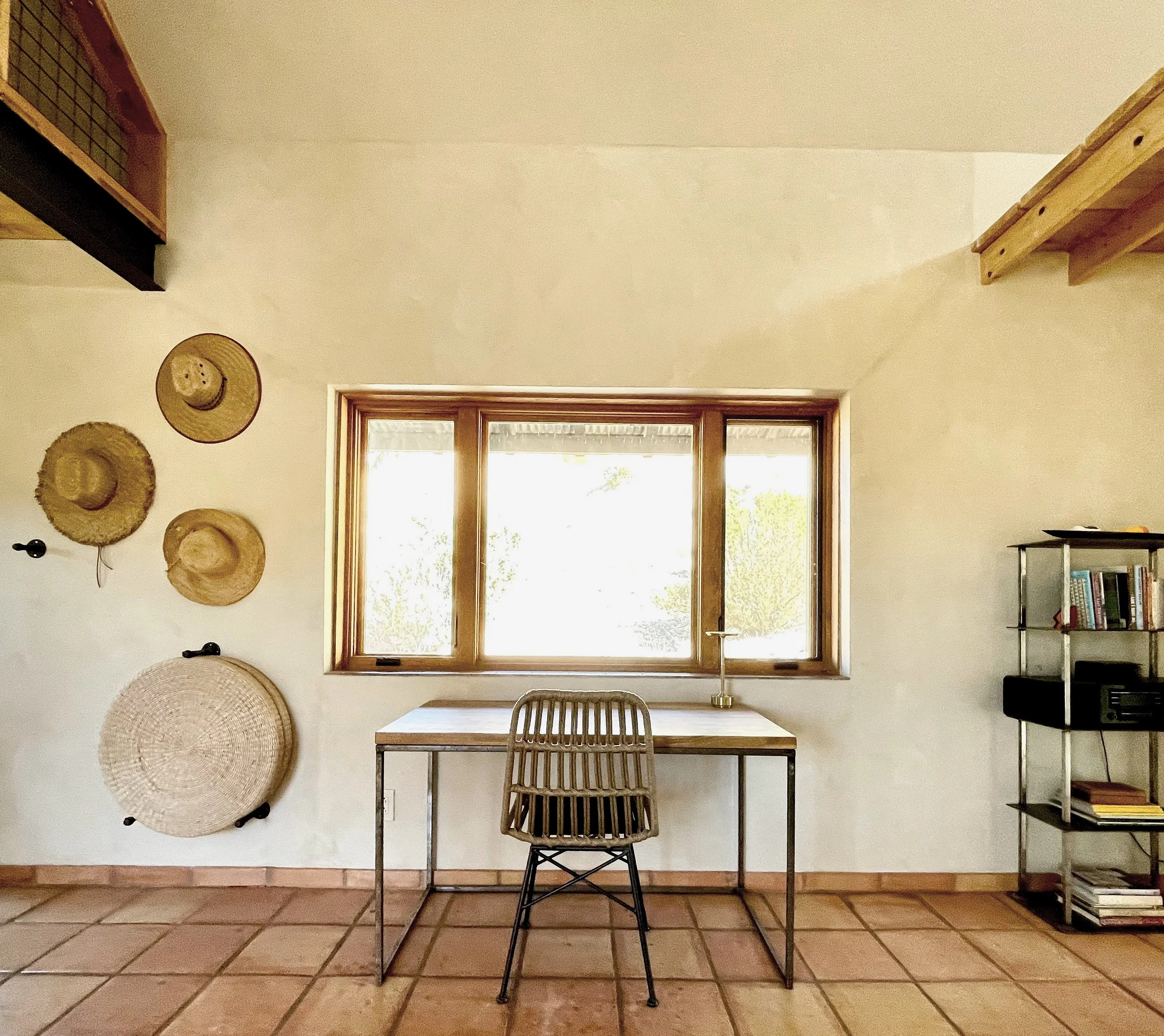 A minimalist room with a wooden desk, a metal-framed chair, a window with wooden framing, and wall decorations including straw hats and woven baskets, with a bookshelf on the right side.