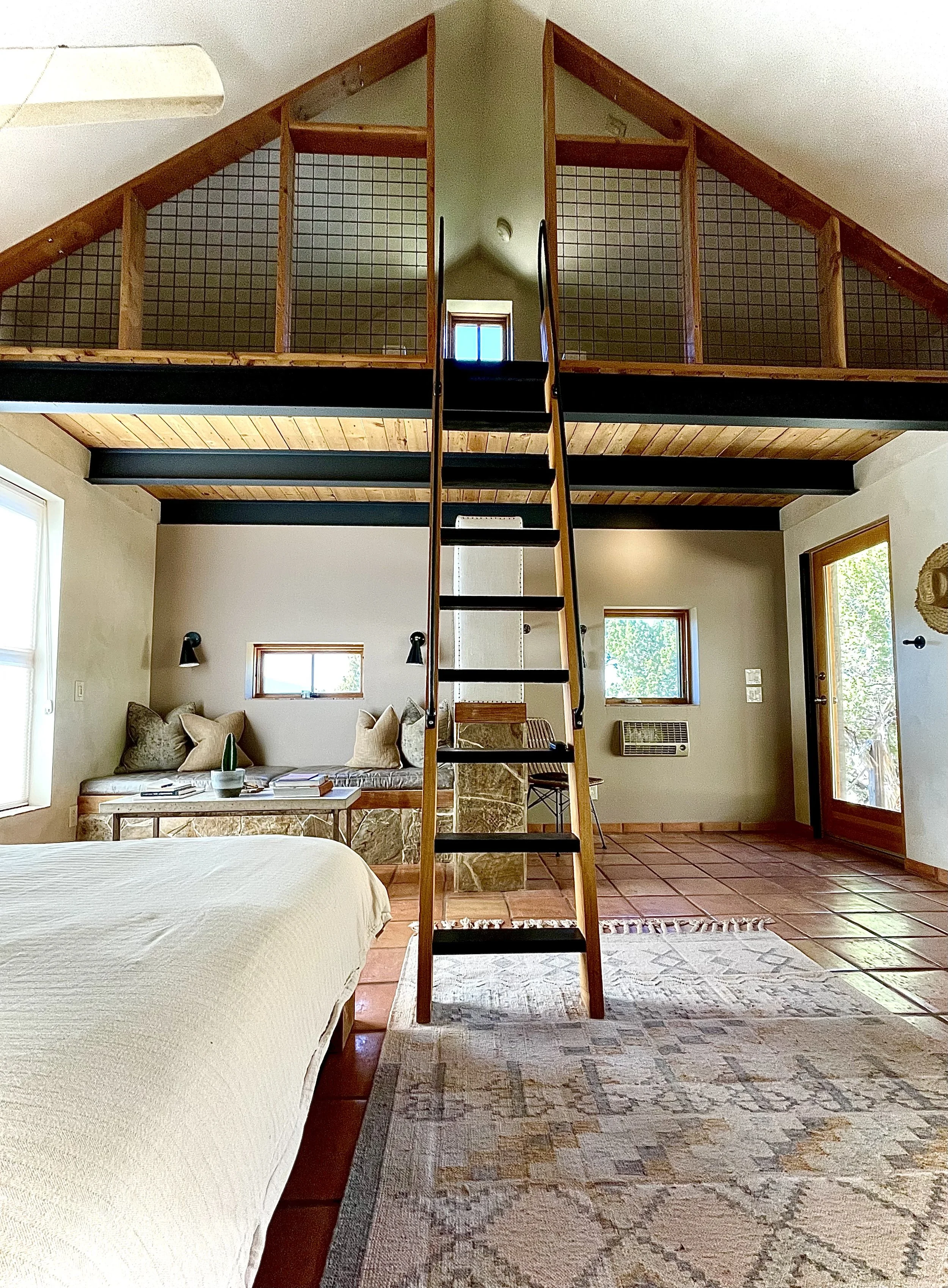 Interior of a bedroom with a lofted area accessed by a ladder, featuring wood and metal railings, two small windows, a bed with white bedding, a stone bench with pillows, and a door leading outside.