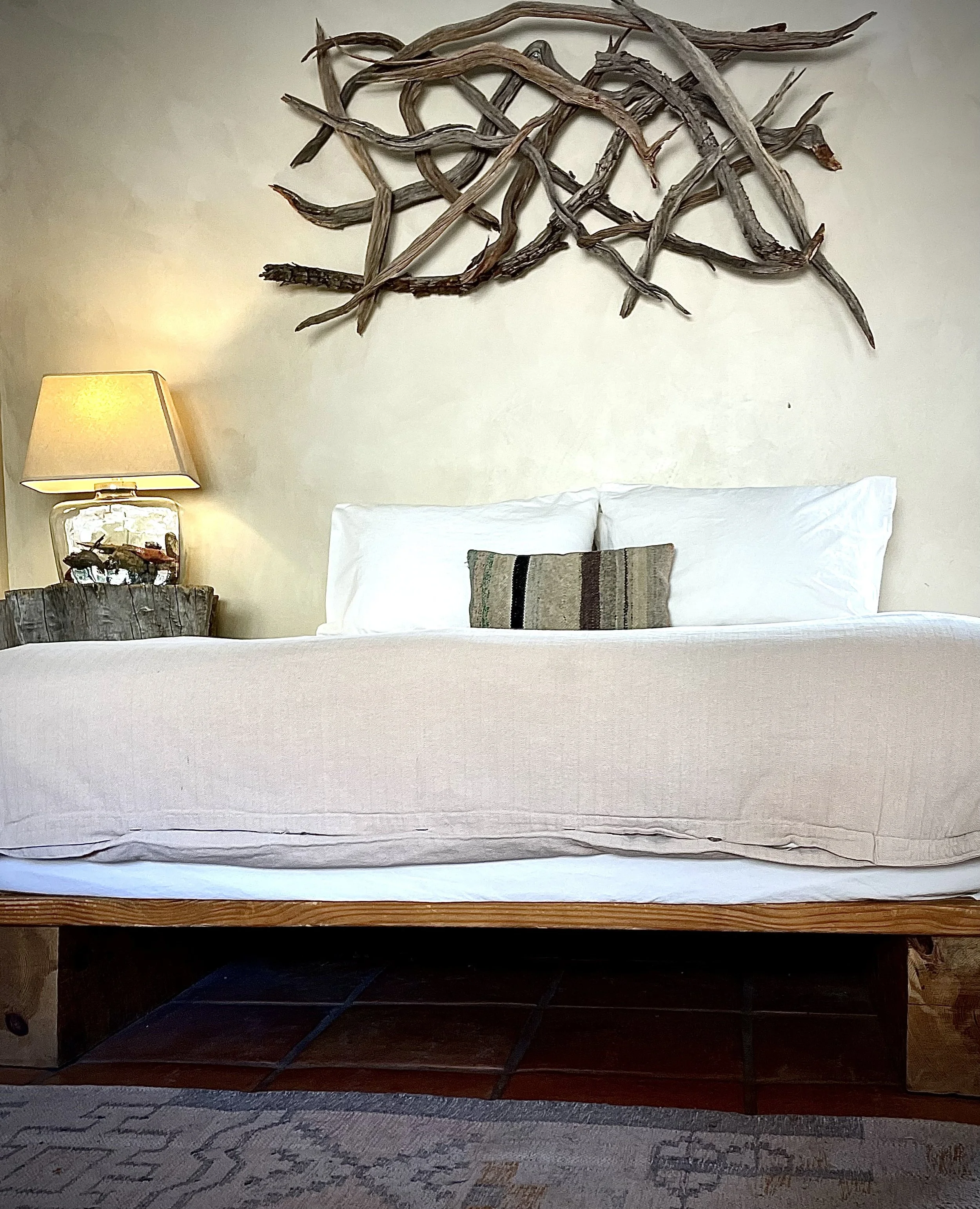 A bedroom with a beige bed and white pillows, above which is a wall decorated with driftwood arranged in a pattern. To the left, there is a lamp on a wooden nightstand.