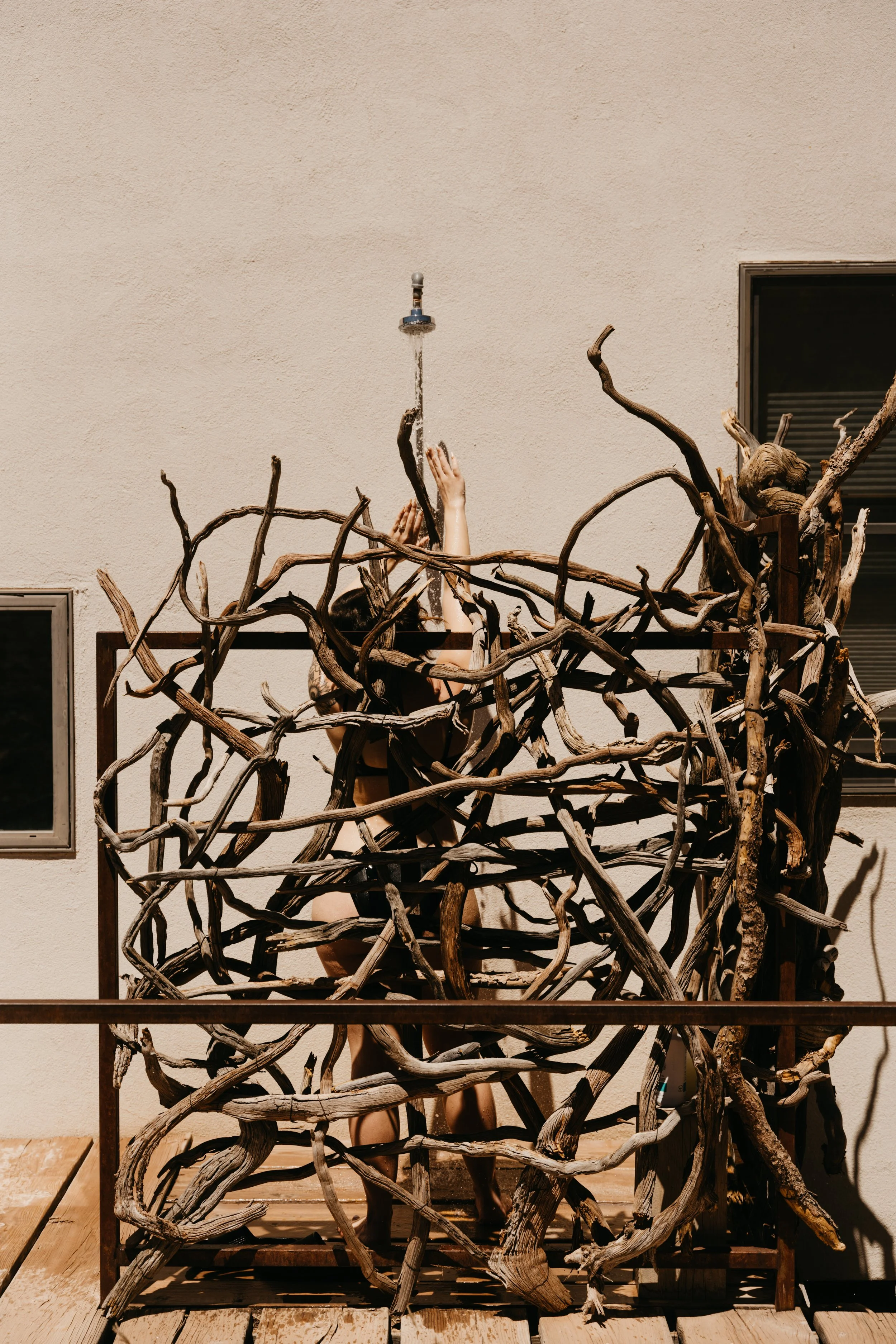 Person standing behind a wooden sculpture with intertwined branches, reaching upward to turn on a water faucet.