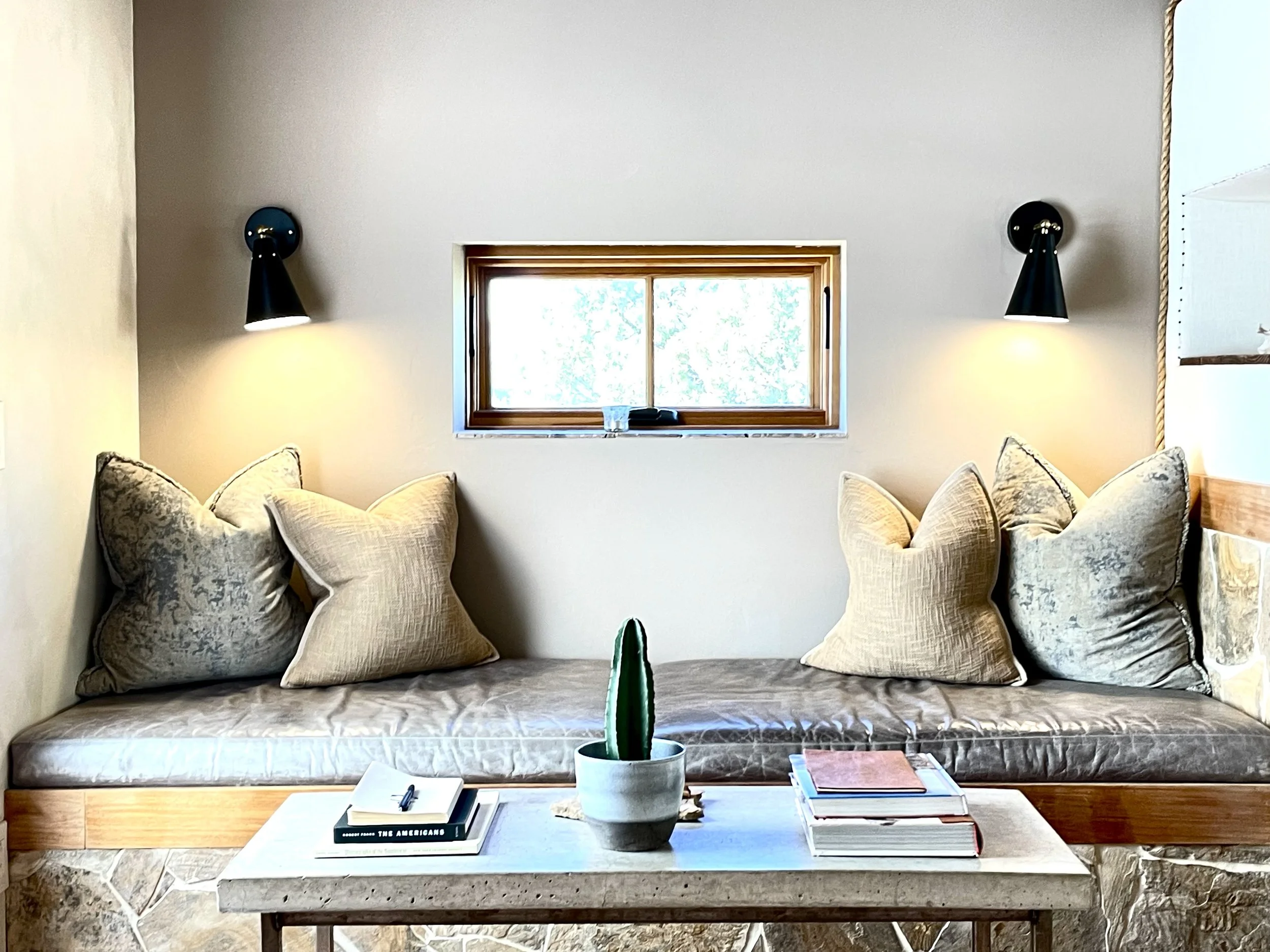 Interior scene of a cozy window seat with cushions, a small table with books and a potted cactus, and wall-mounted lamps on either side of the window.