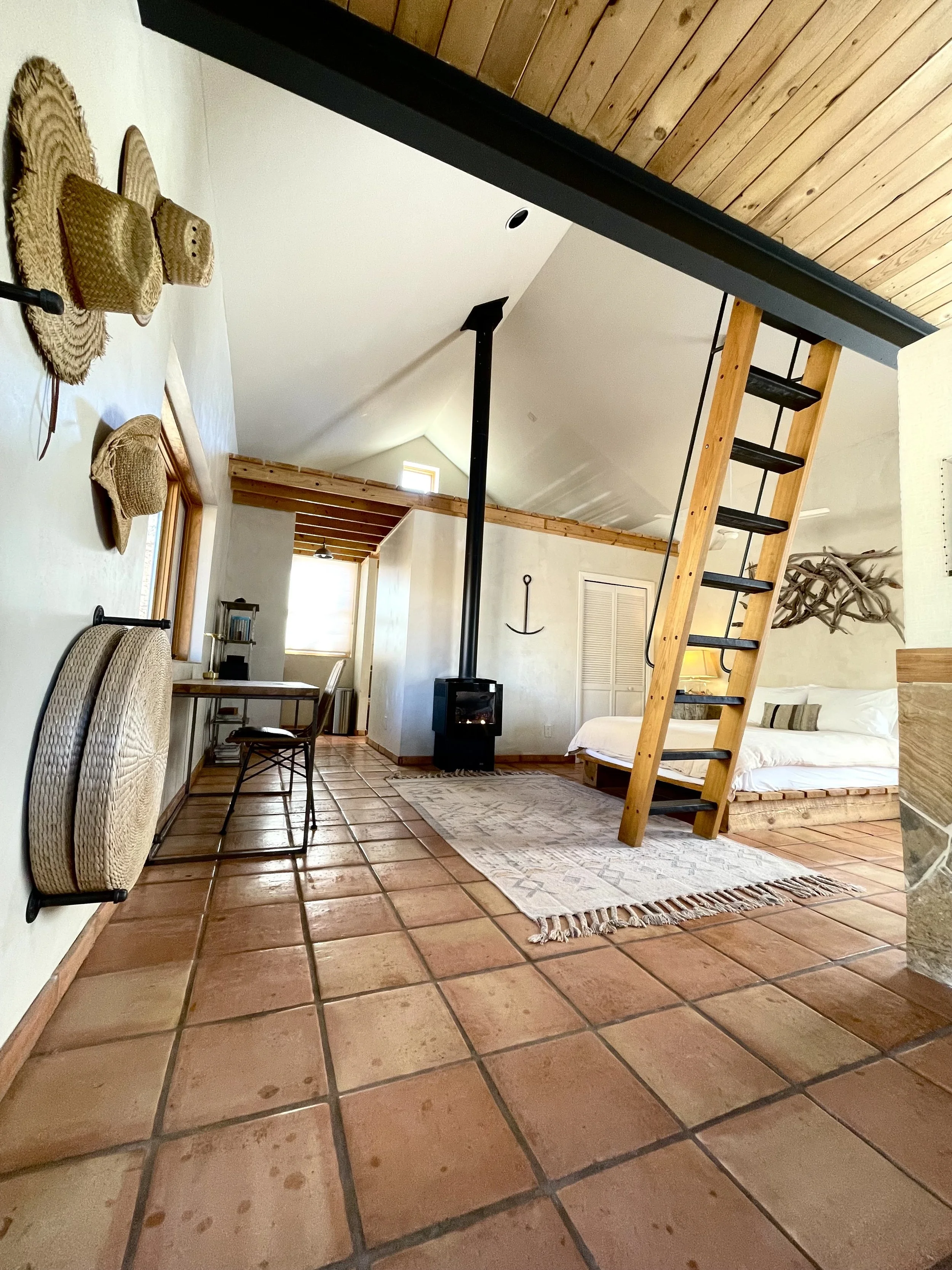 Interior of a rustic-style room with terracotta tile flooring, a sloped ceiling with wood paneling, a wood staircase leading to a loft, a white bed with a wooden headboard, a small black wood stove, and wall decor including hats, a framed picture, and a wall hanging.