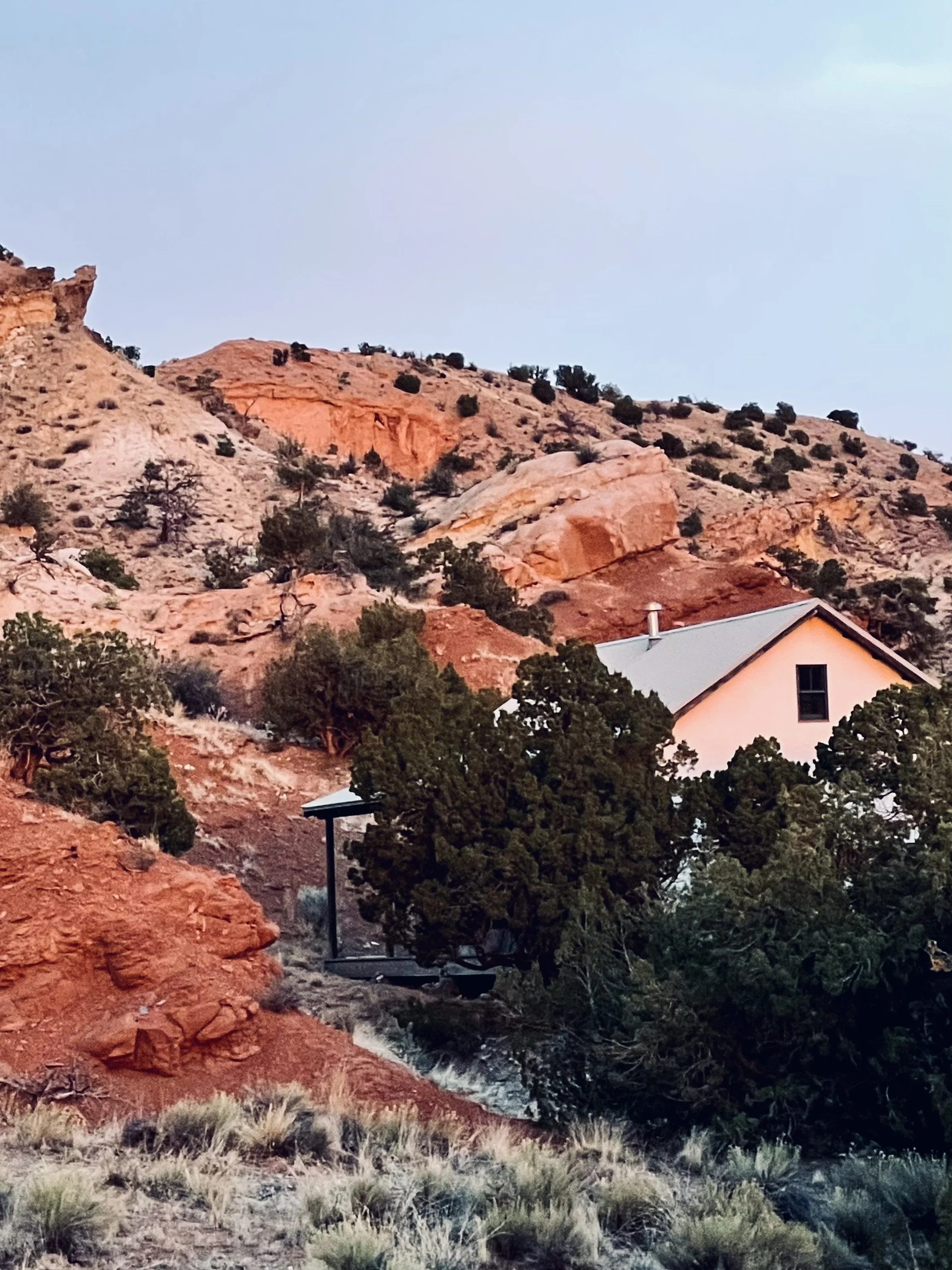 A house with a gray roof and white walls situated among green trees on a slope with reddish rocky hills in the background during twilight.