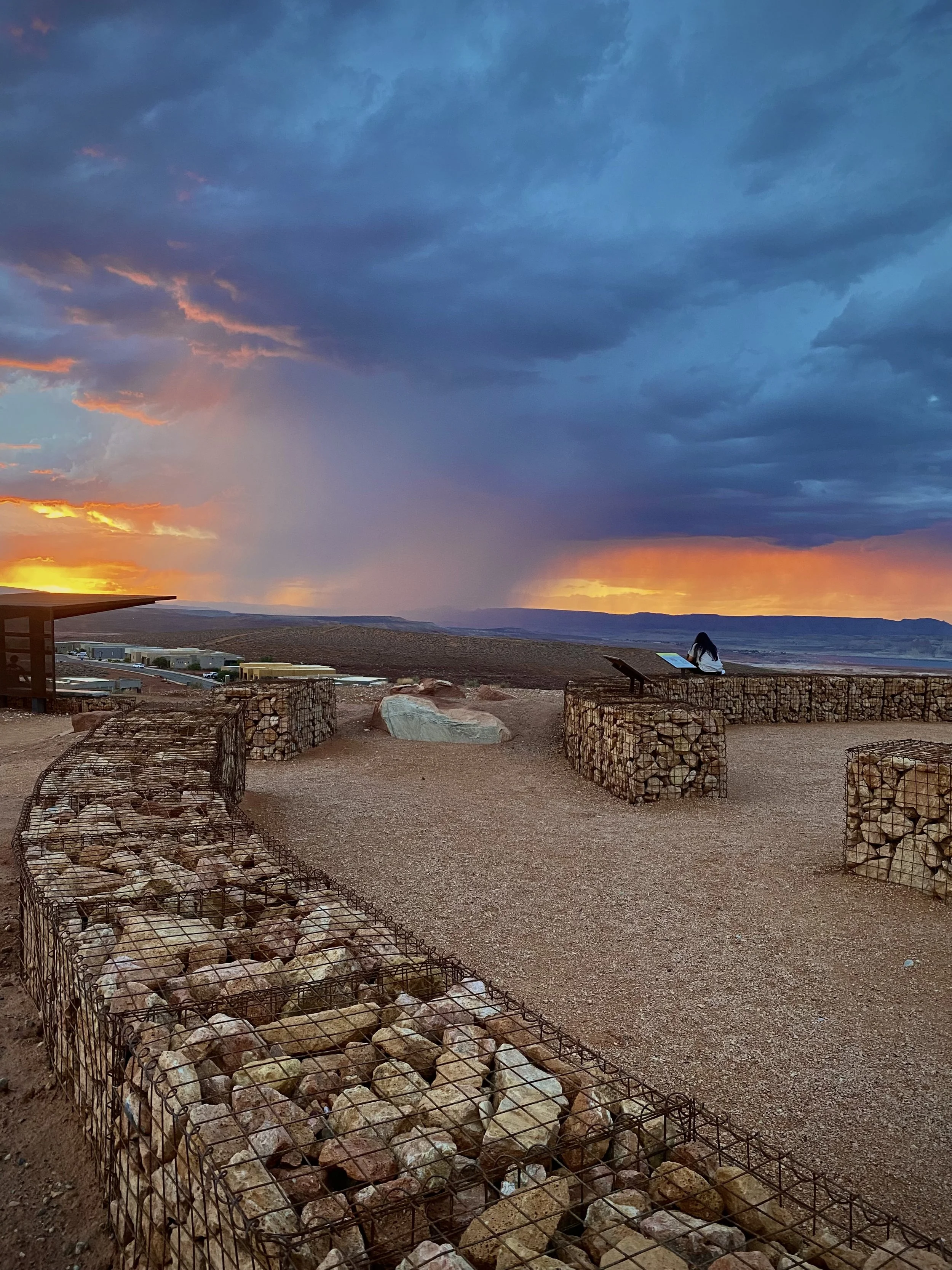 Person sitting on a wall at Grandview Knoll Overlook with distant rainstorm at sunset