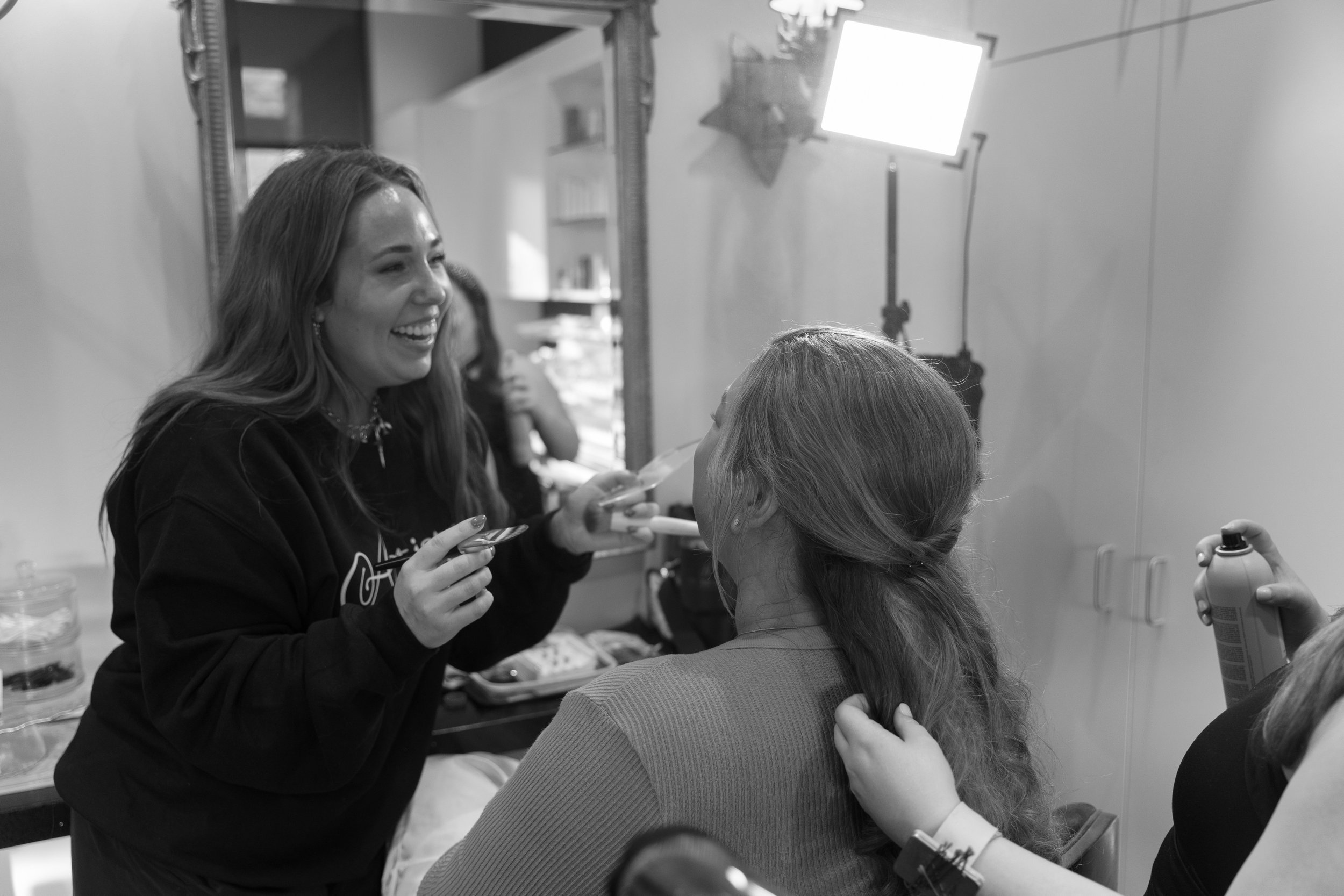 Woman with styled red hair having makeup applied by a makeup artist, with others in the background.