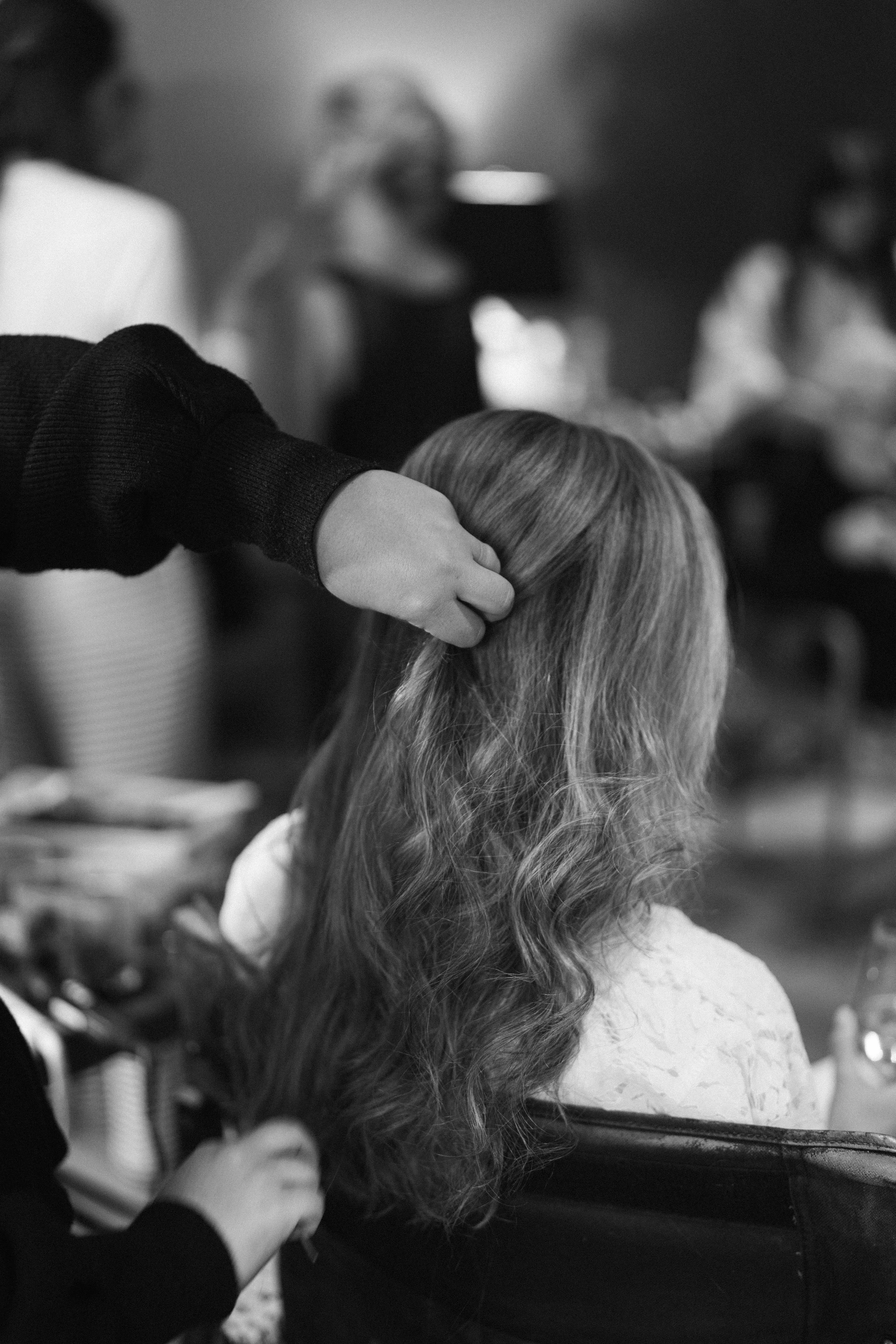 A woman with styled blonde hair and pearl hairpins getting makeup done by a makeup artist with a mirror in hand in a bright room.