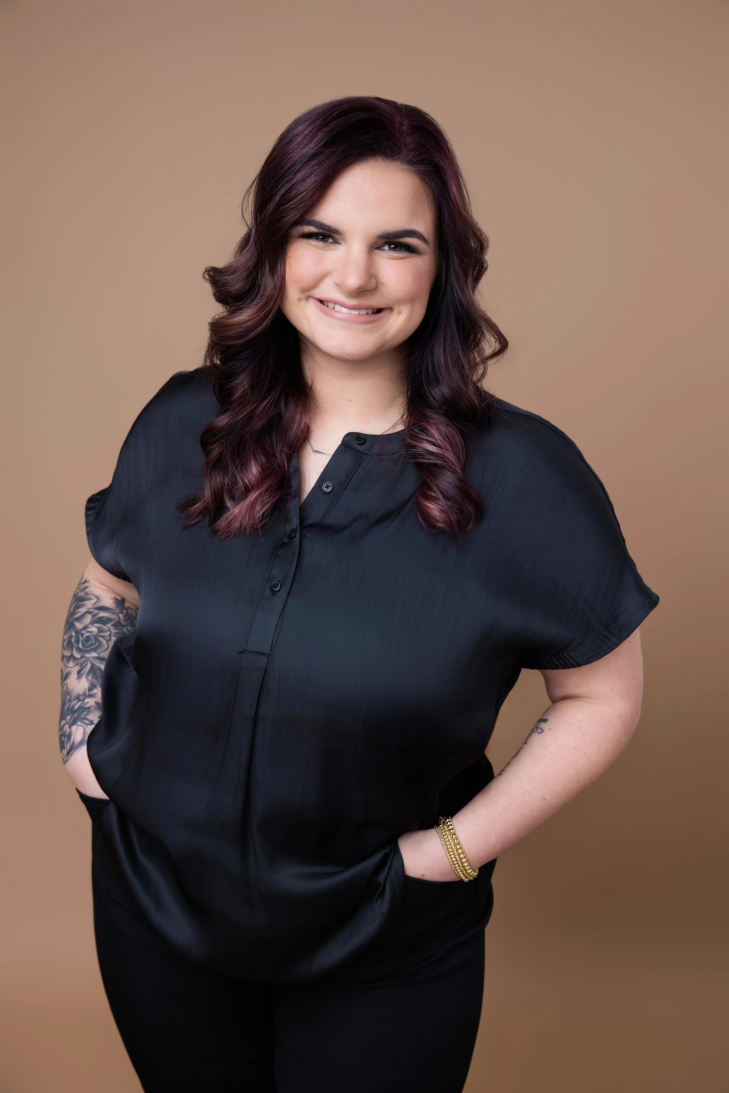 A young woman with dark purple hair smiling, wearing a black blouse, standing against a beige background.