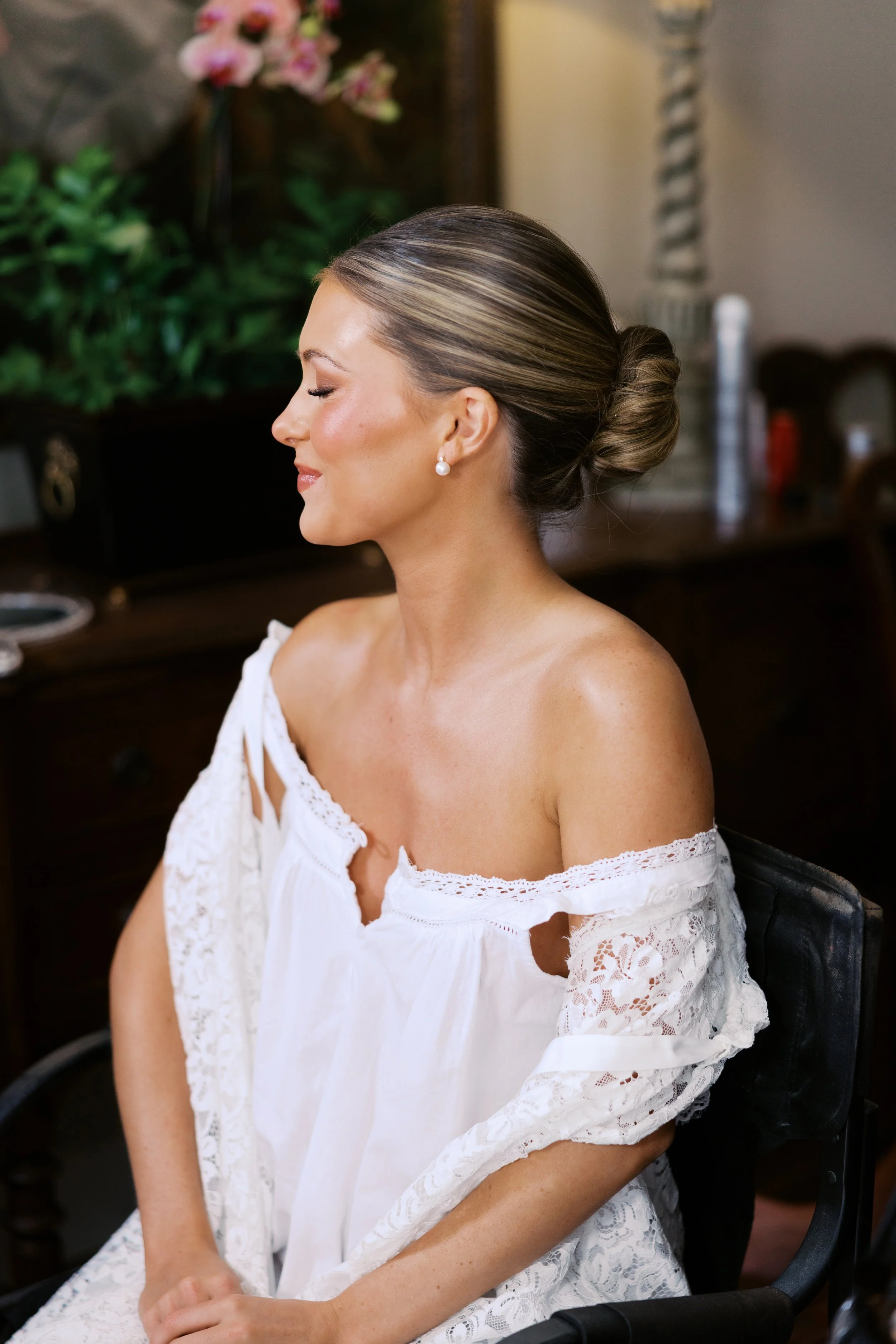 Bridal portrait of a woman in a white wedding dress with lace detailing, wearing jewelry including earrings, necklace, bracelet, and wristwatch, and holding a white veil with an outdoor urban background.