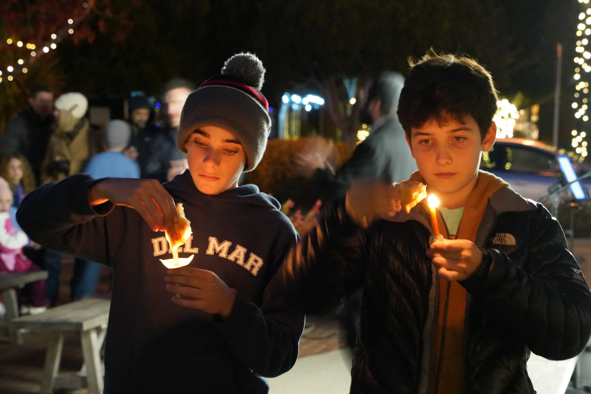 Two boys holding lit candles at night during a festive outdoor event, with people and holiday lights in the background.