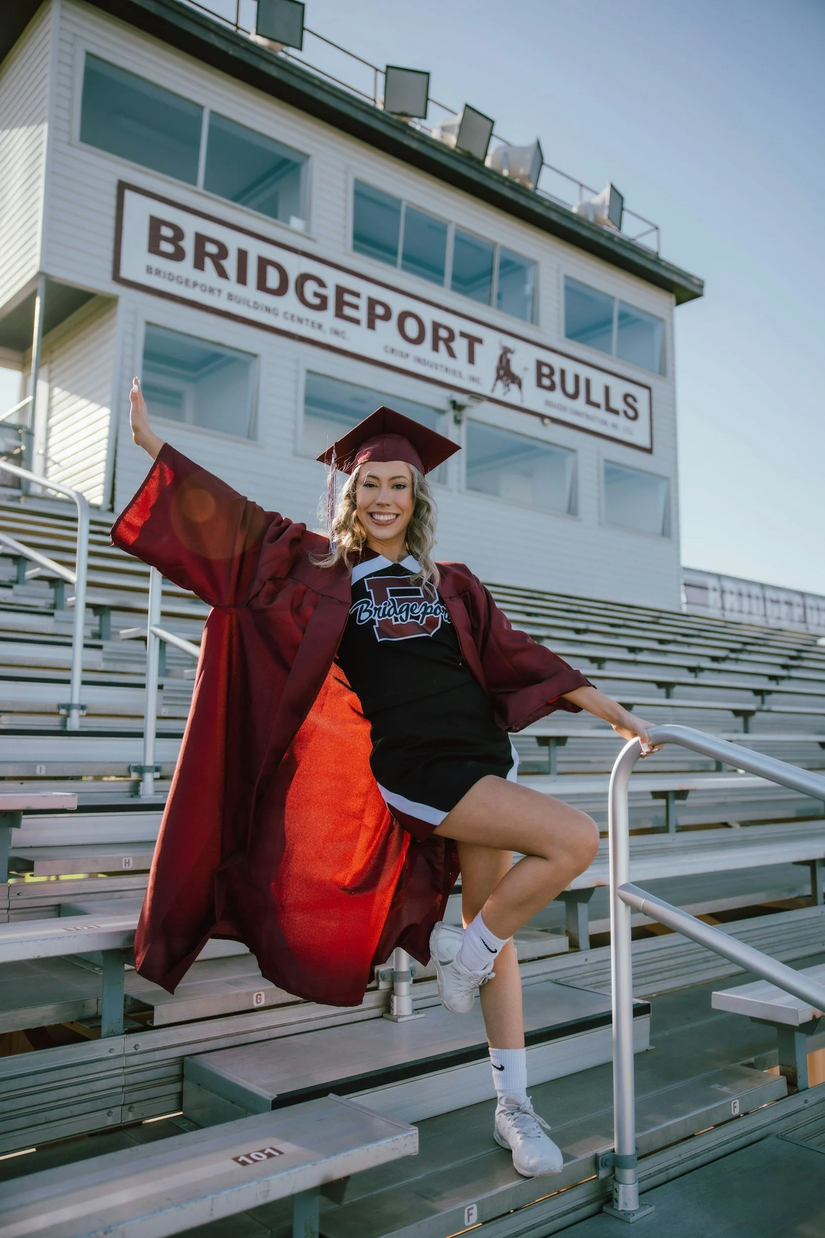 A smiling young woman in a graduation cap and gown raising her right arm, sitting on stadium bleachers with a large sign that says 'Bridgeport Bulls' in the background.
