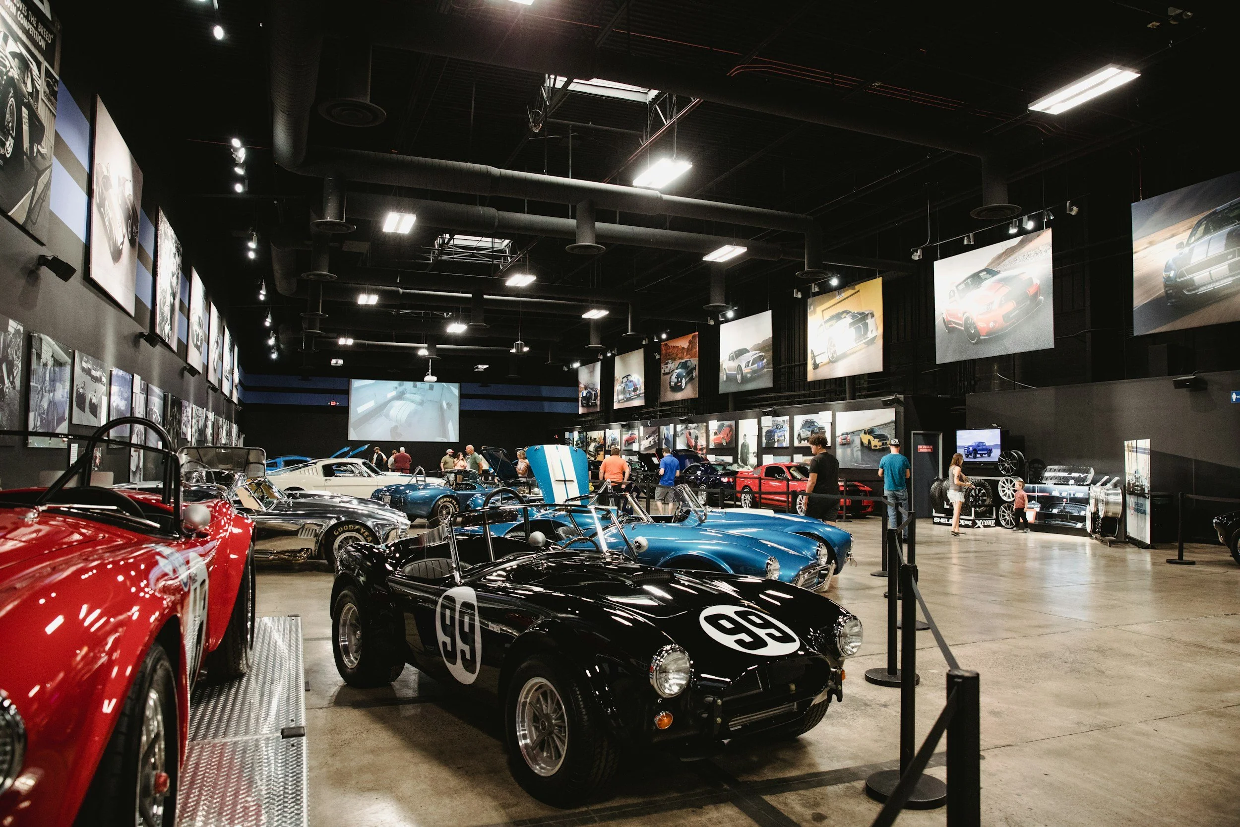 Indoor display of classic and vintage cars at an automobile museum, with pictures of cars on the walls and visitors walking around.