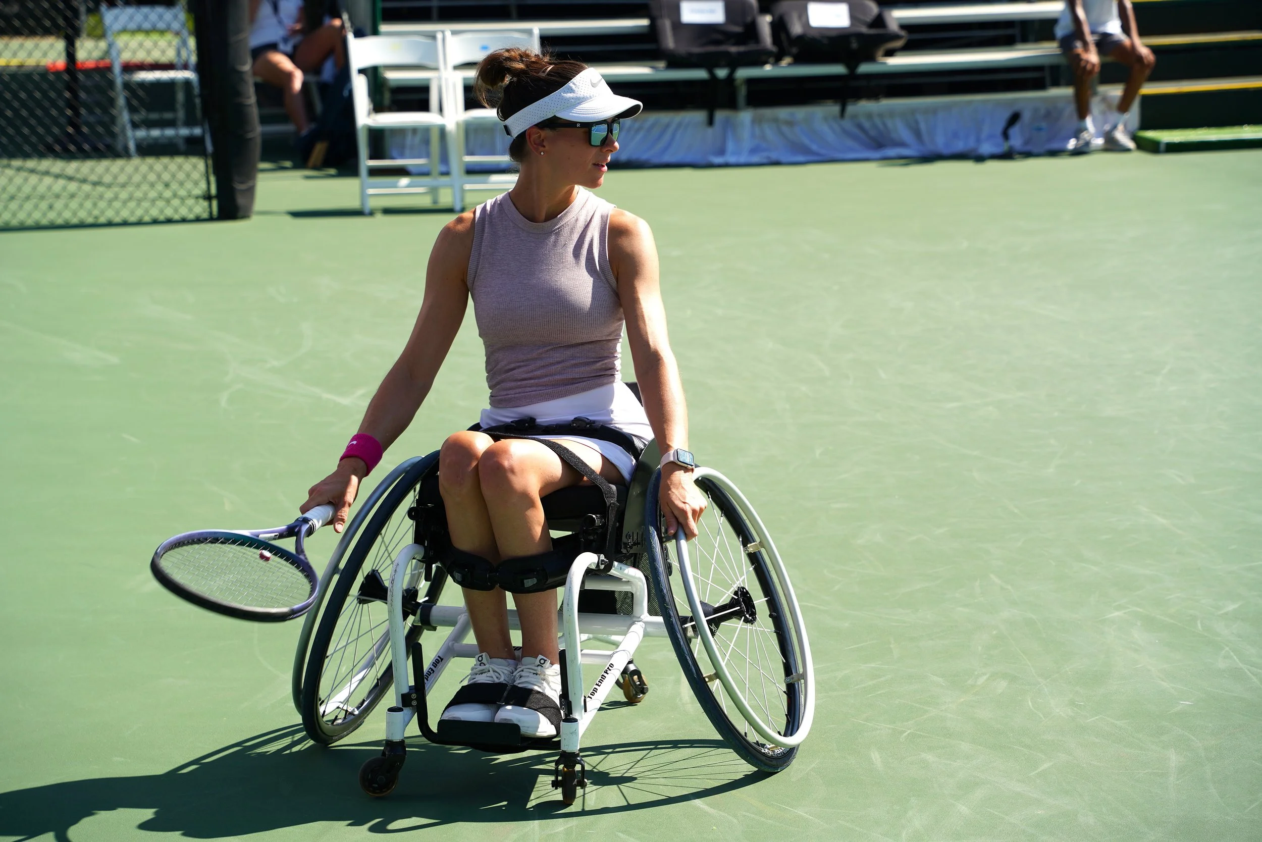 A woman with sunglasses and a visor in a wheelchair holding a tennis racket on a tennis court.