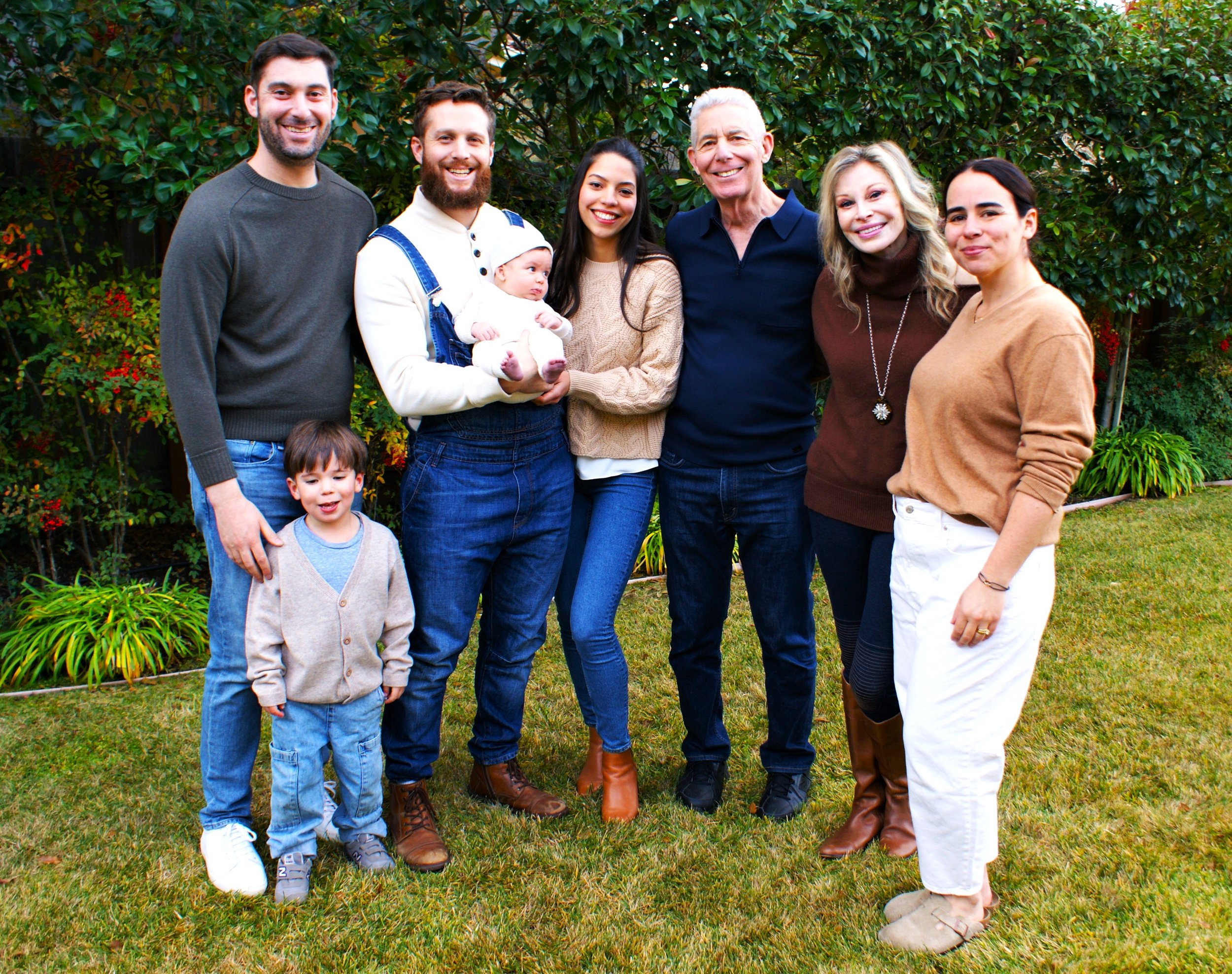 A diverse group of eight people, including children and adults, standing outdoors on grass in front of dense green shrubs, smiling, with some holding children.