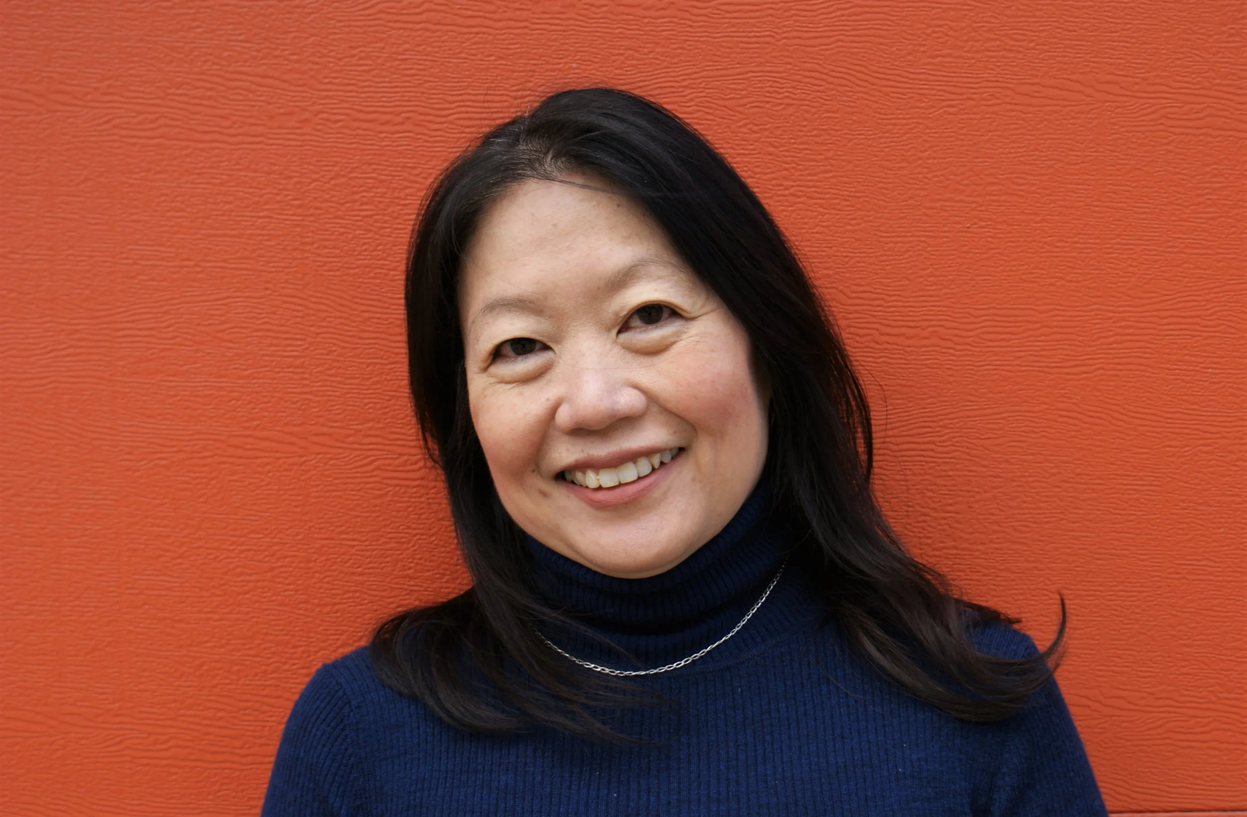 A woman with long black hair, fair skin, wearing a dark blue turtleneck sweater and a necklace, smiling, standing in front of an orange textured wall.