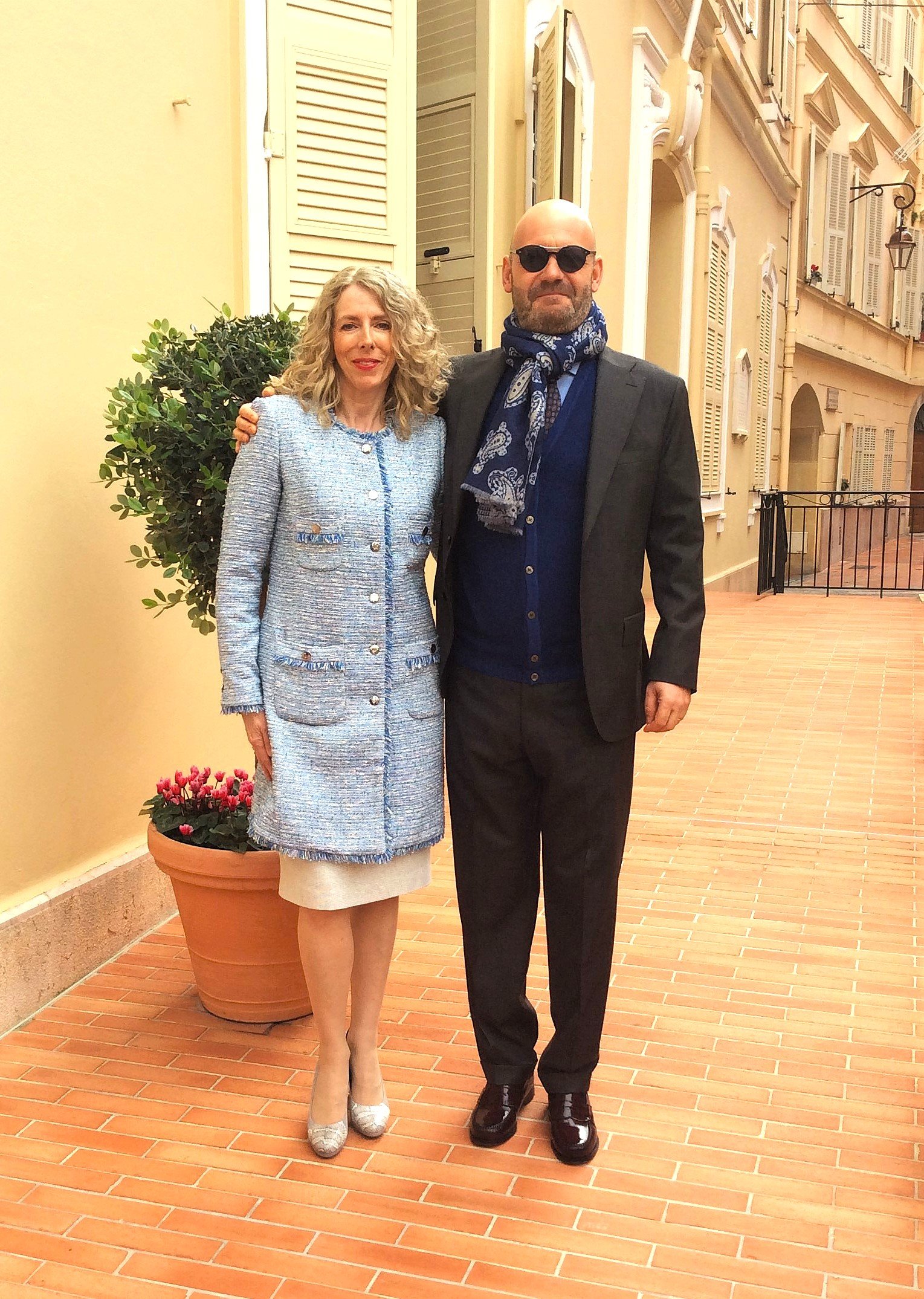 A woman and a man standing side by side outdoors, smiling at the camera. The woman is wearing a light blue daytime suit with a white skirt, beige heels, and has curly blonde hair. The man is dressed in a black suit with a blue shirt, a patterned scarf, sunglasses, and black shiny shoes. They are standing on a terracotta-tiled sidewalk next to a potted plant with pink flowers and a yellow building with shutters and decorative window frames in the background.