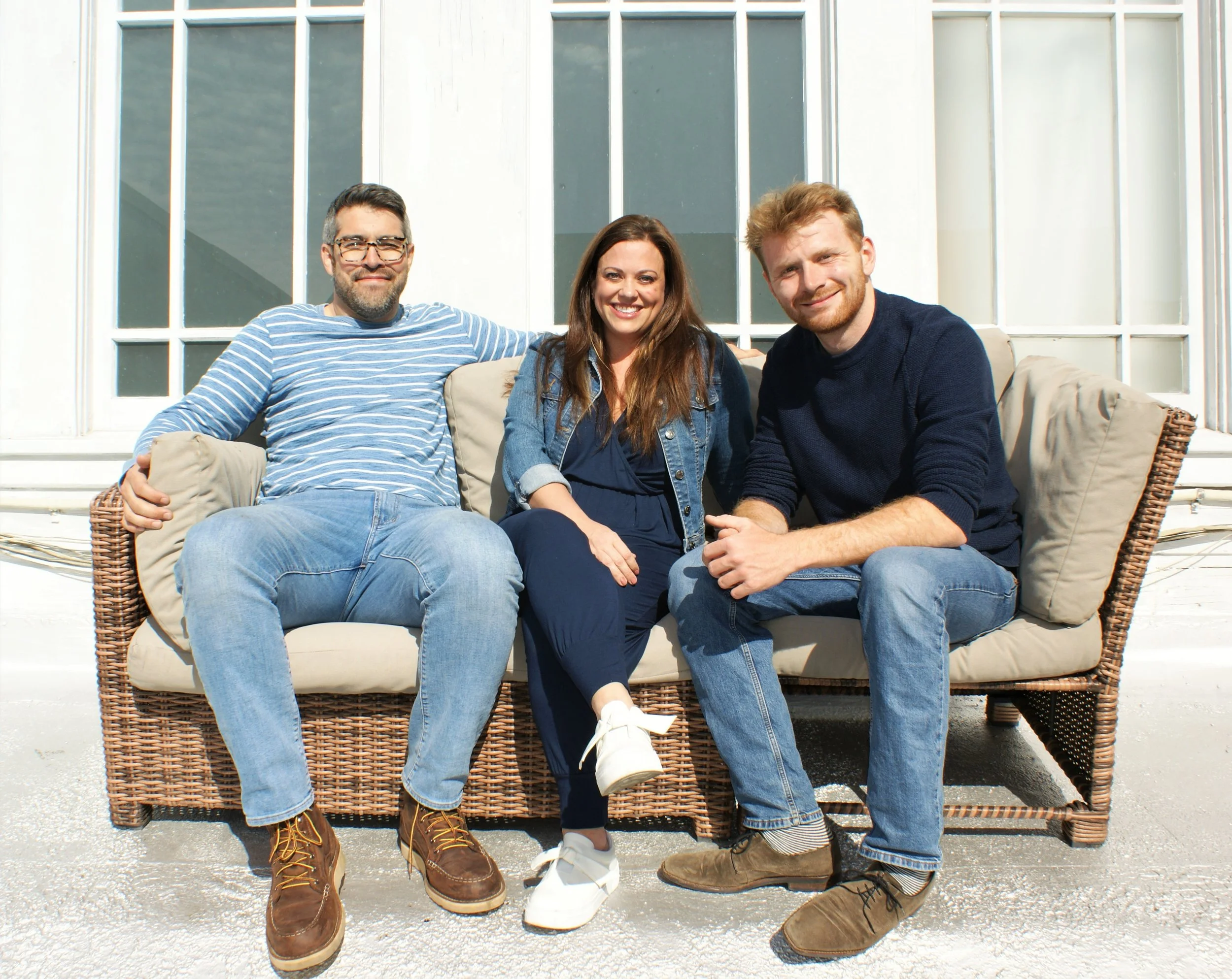 Three friends sitting on a wicker outdoor sofa in front of a large window, smiling at the camera on a sunny day.