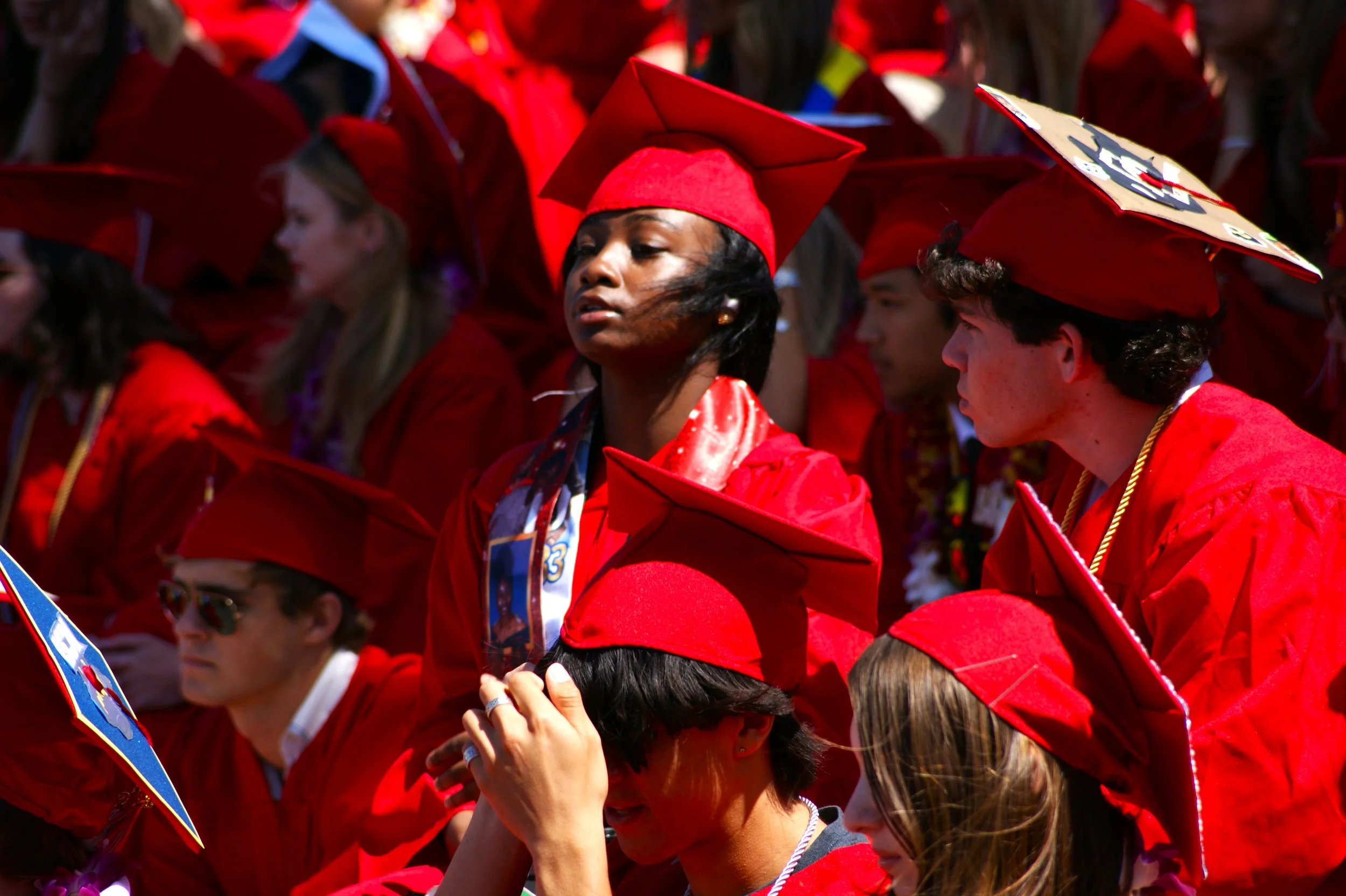 Many graduates in red caps and gowns at a graduation ceremony, with some wearing medals or cords, outdoors.