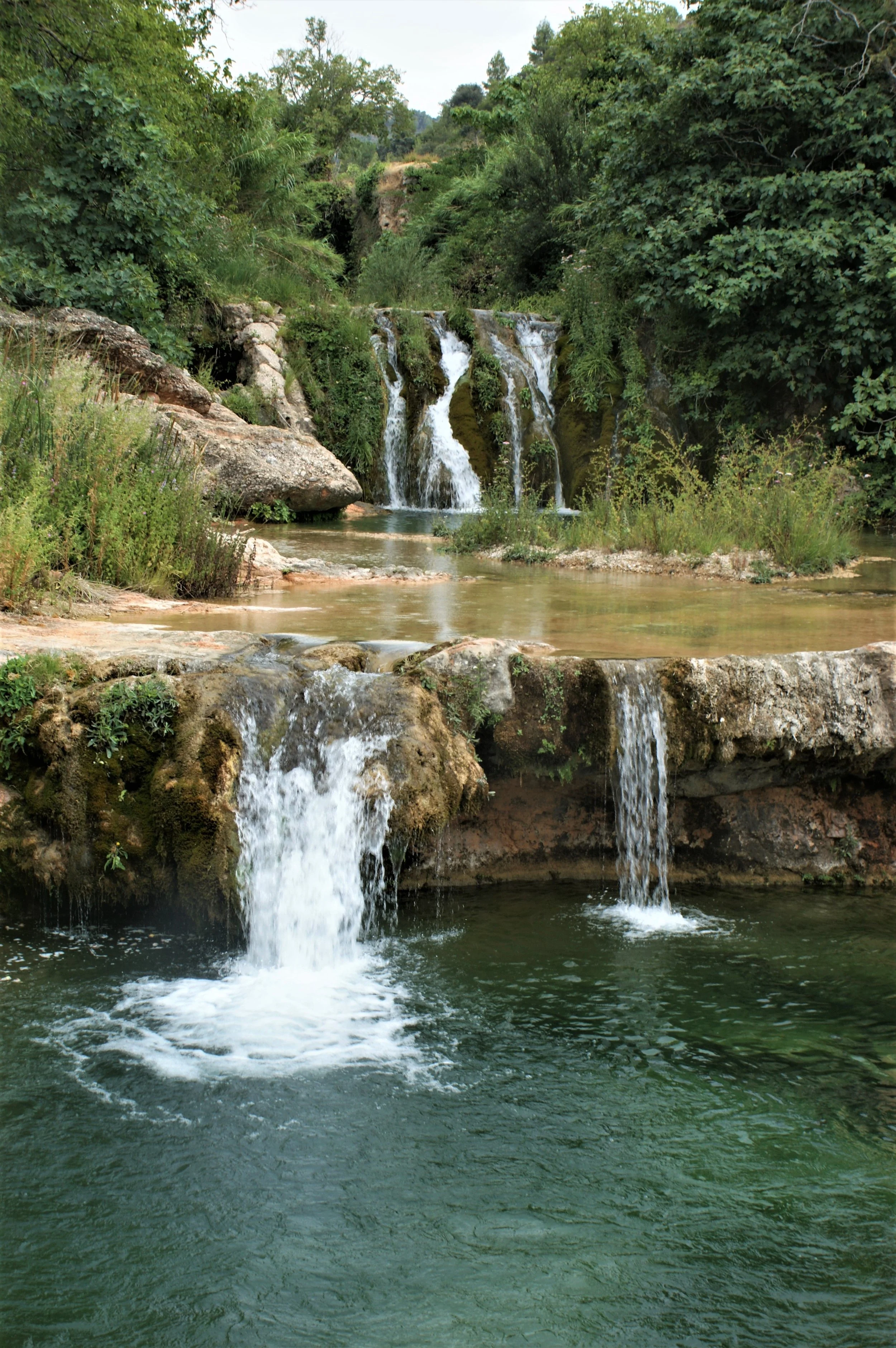 Small waterfall flowing into a clear pond surrounded by green trees and bushes.