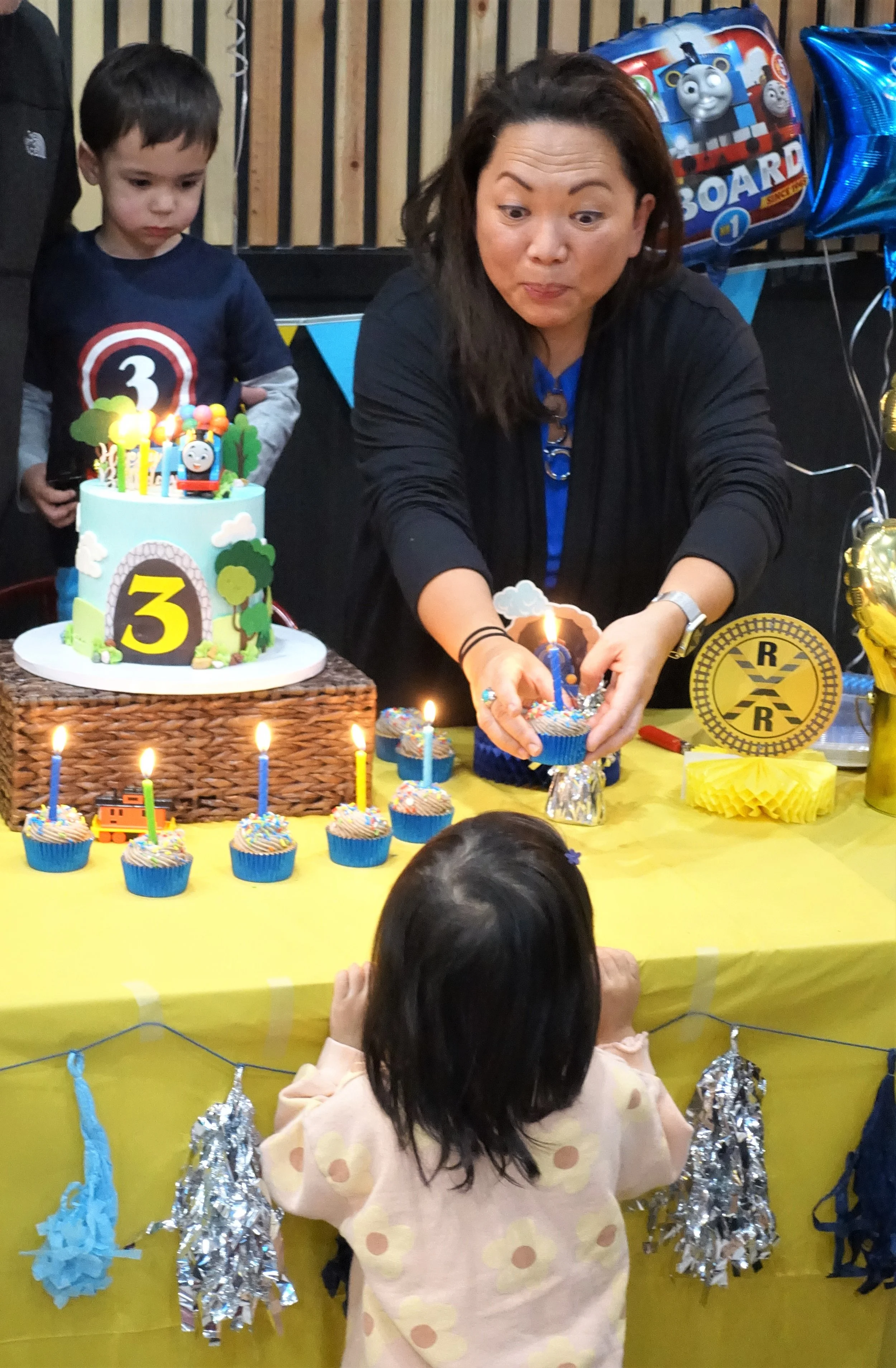 A birthday party scene with children and an adult. There are cakes, cupcakes, and balloons decorated for a celebration. A young boy looks at a cake with a number 3 on it, and an adult woman is placing candles on a cupcake.