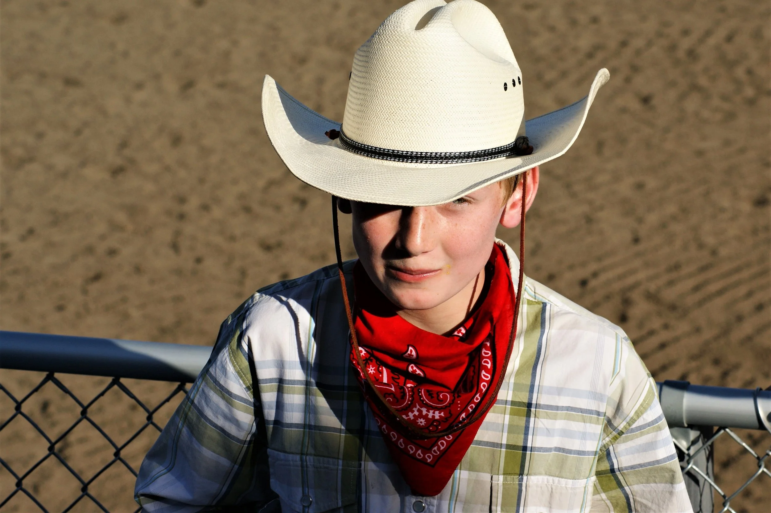 Young person wearing a large white cowboy hat with black accents, a red bandana around their neck, and plaid shirt, standing behind a chain-link fence with a sandy area in the background.
