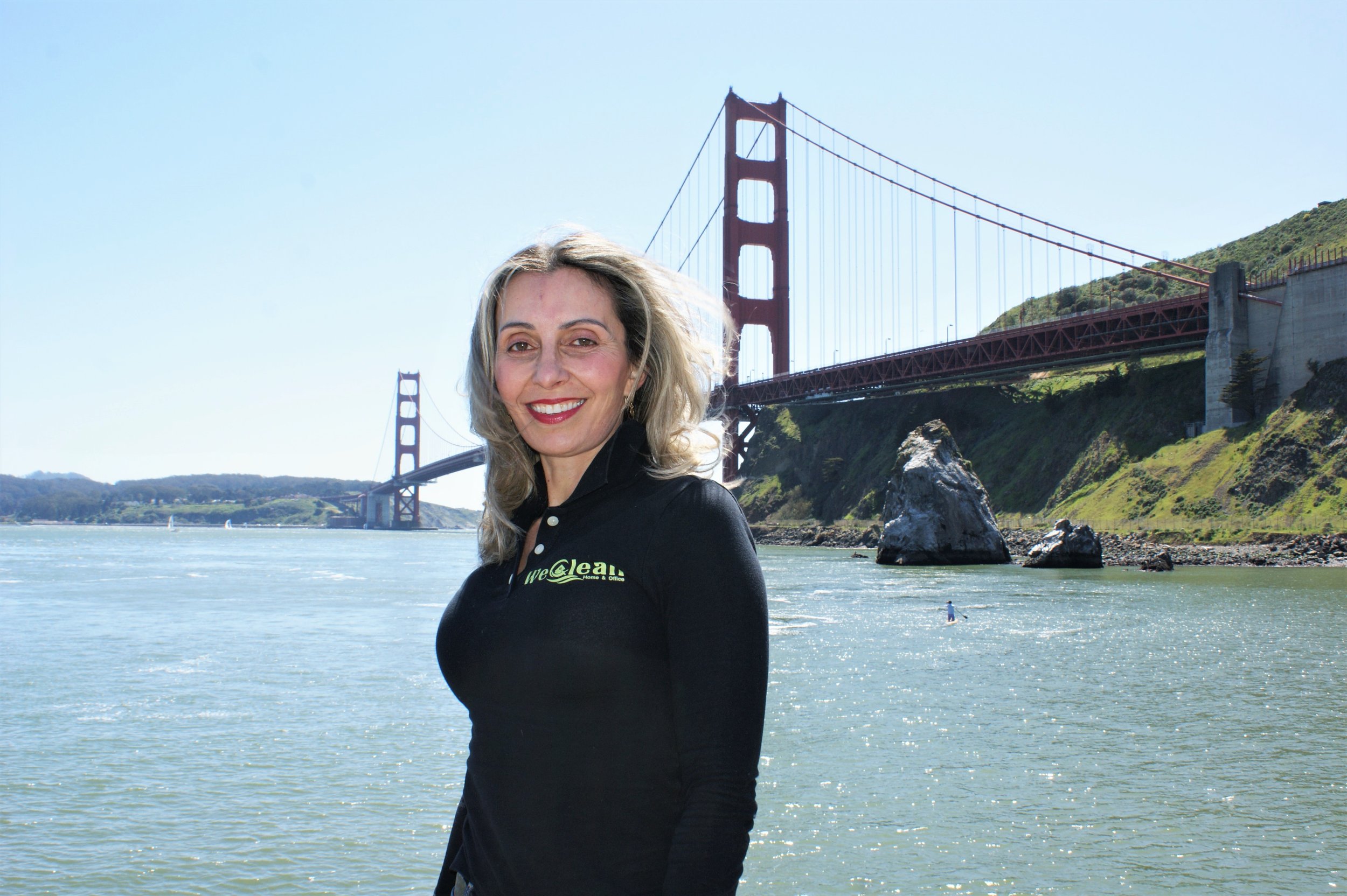 A woman with blonde hair smiling outdoors near the water with the Golden Gate Bridge in the background.